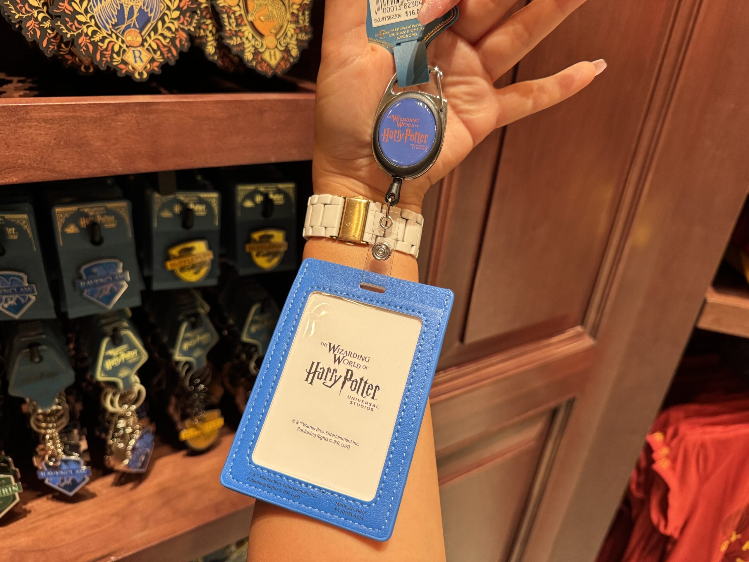 A hand holds a blue Harry Potter lanyard badge holder in a gift shop filled with Wizarding World merchandise.
