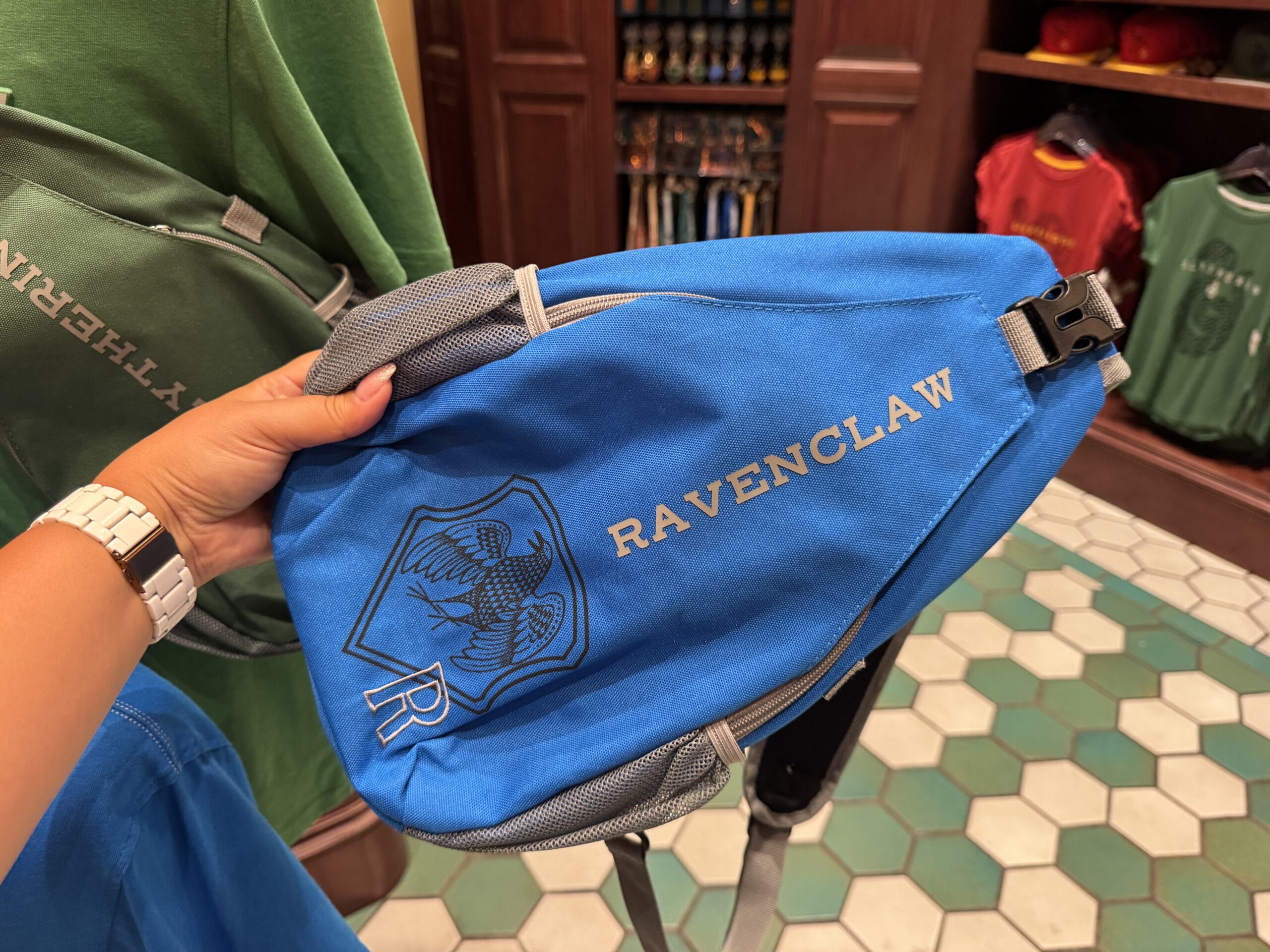 A hand holds a blue Ravenclaw sling bag, Epic Universe Wizarding World of Harry Potter merchandise, in a store background.