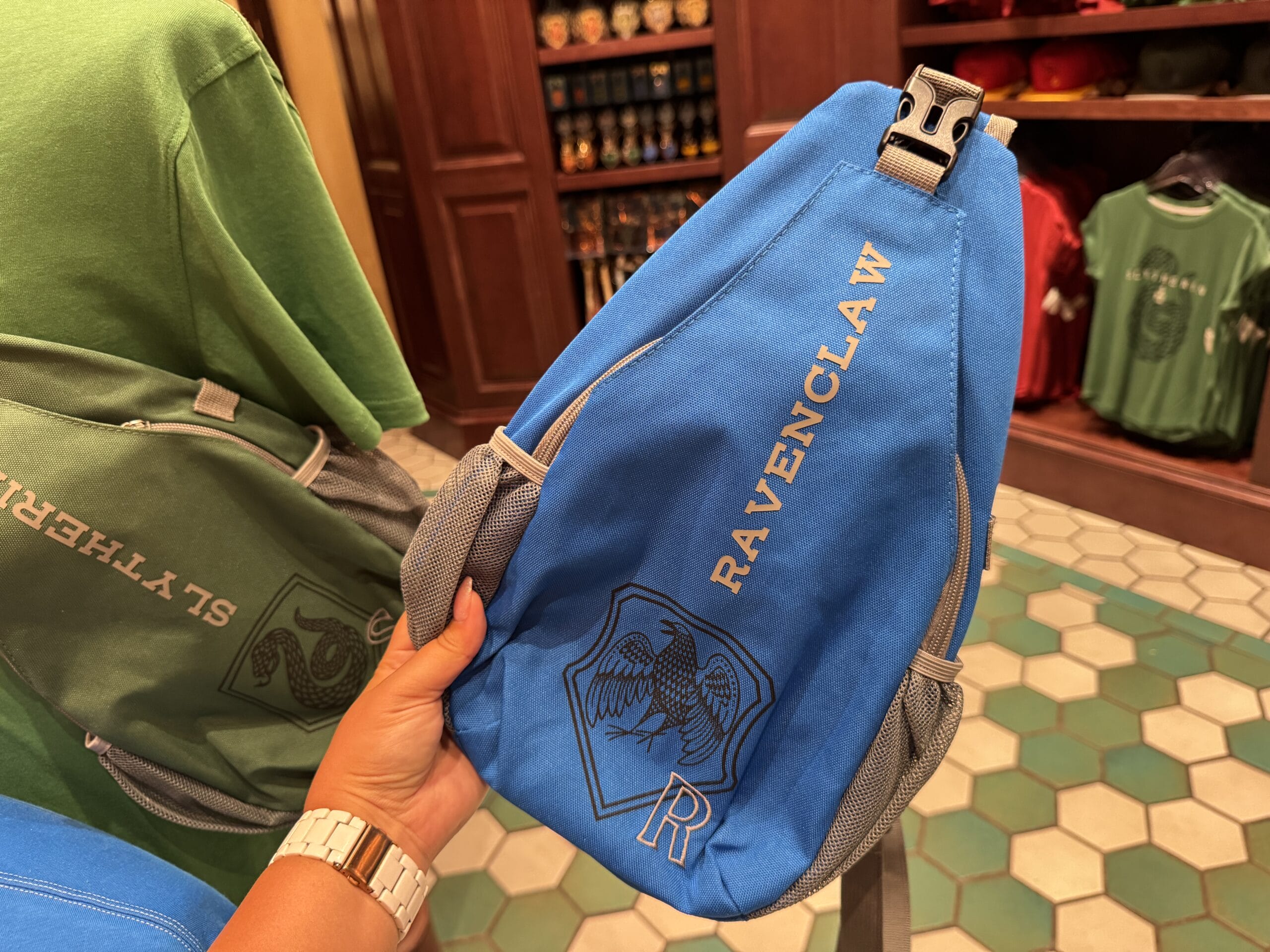 Hand holding a blue Ravenclaw sling bag with house crest; Epic Universe Wizarding World of Harry Potter merchandise. Slytherin shirt in back.