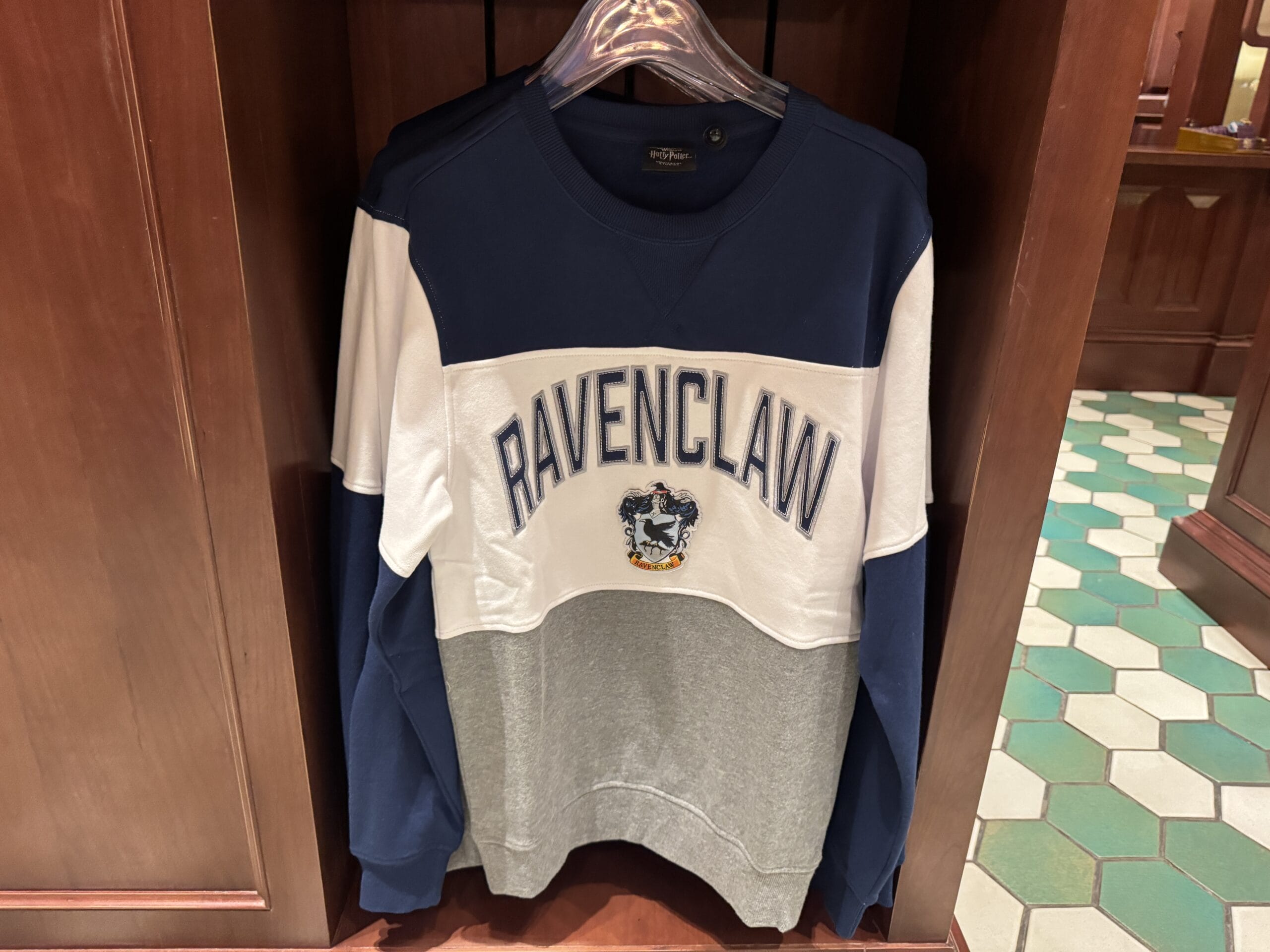 A Ravenclaw sweatshirt with blue, white, and gray panels hangs on a rack of Wizarding World merchandise.
