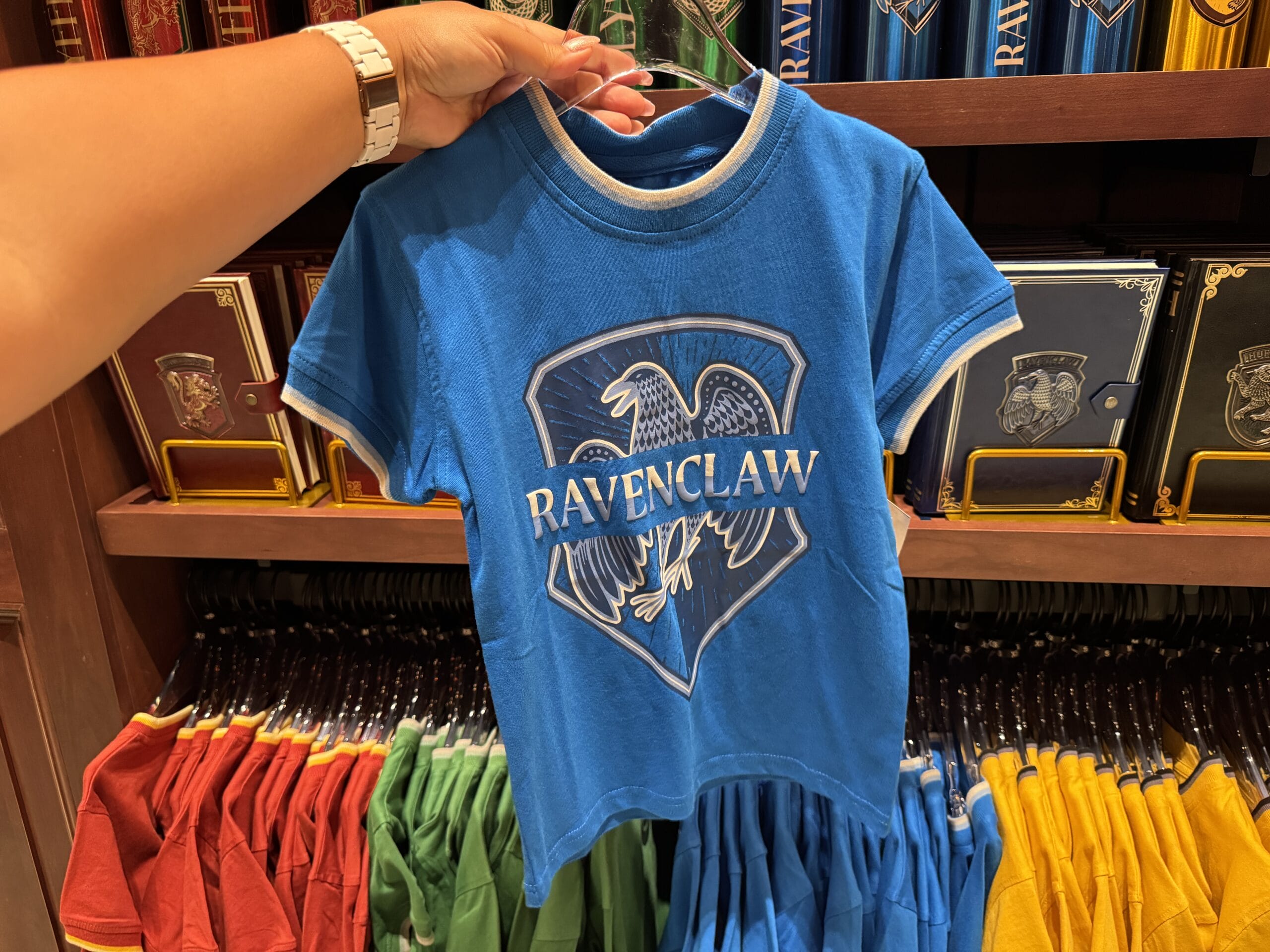 A hand holds a blue Ravenclaw T-shirt, part of Wizarding World of Harry Potter merchandise at Epic Universe.