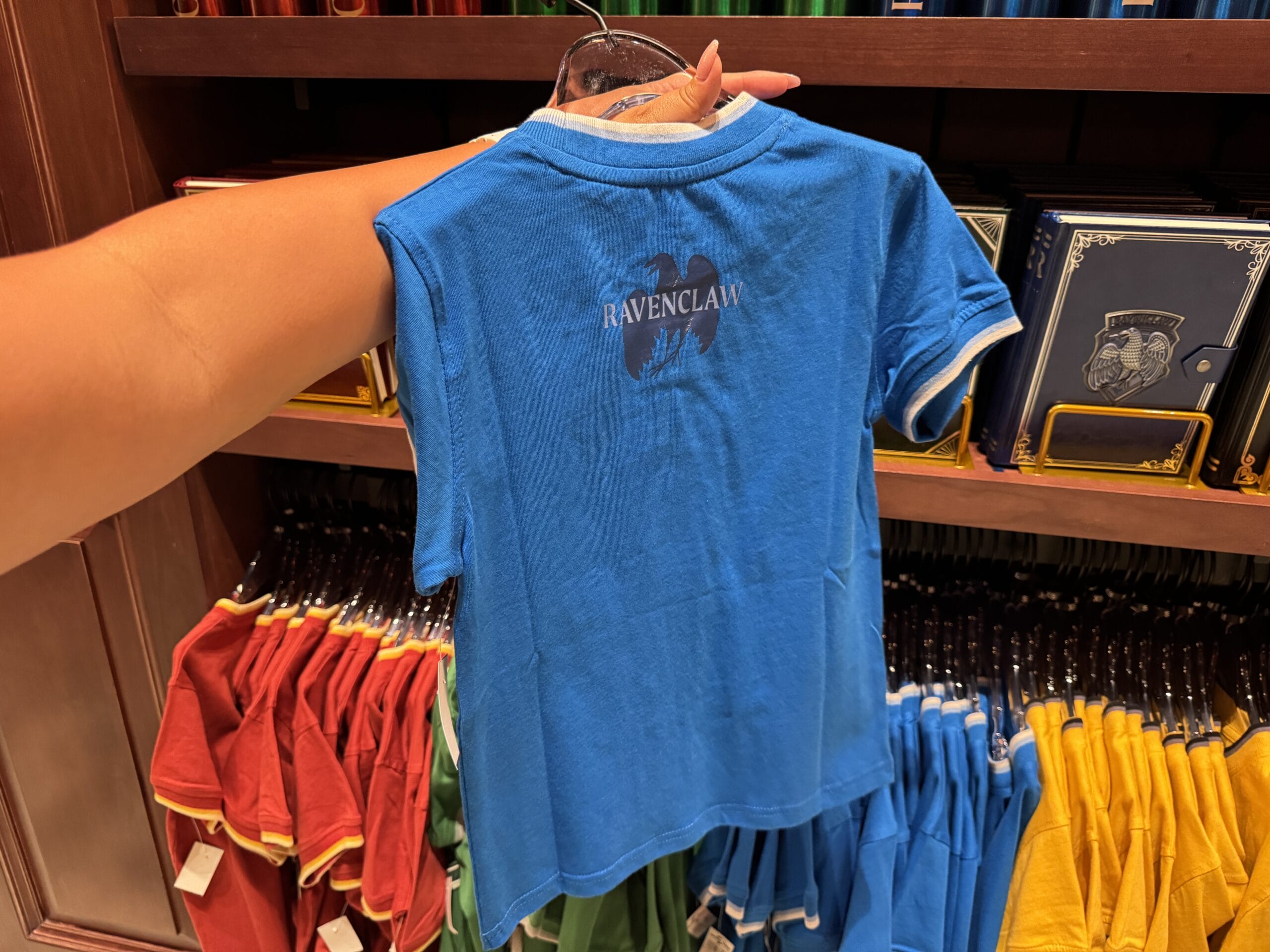 A hand holds up a blue Ravenclaw shirt—Wizarding World of Harry Potter merchandise—amid colorful shirts below.