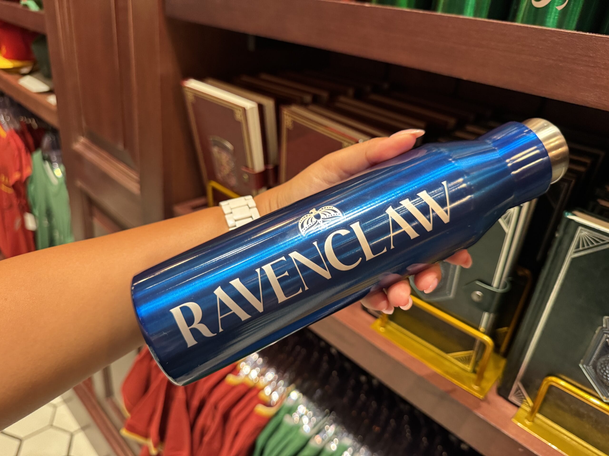 A hand holds a blue "RAVENCLAW" water bottle—magical Harry Potter merchandise among shelves of books and clothes.