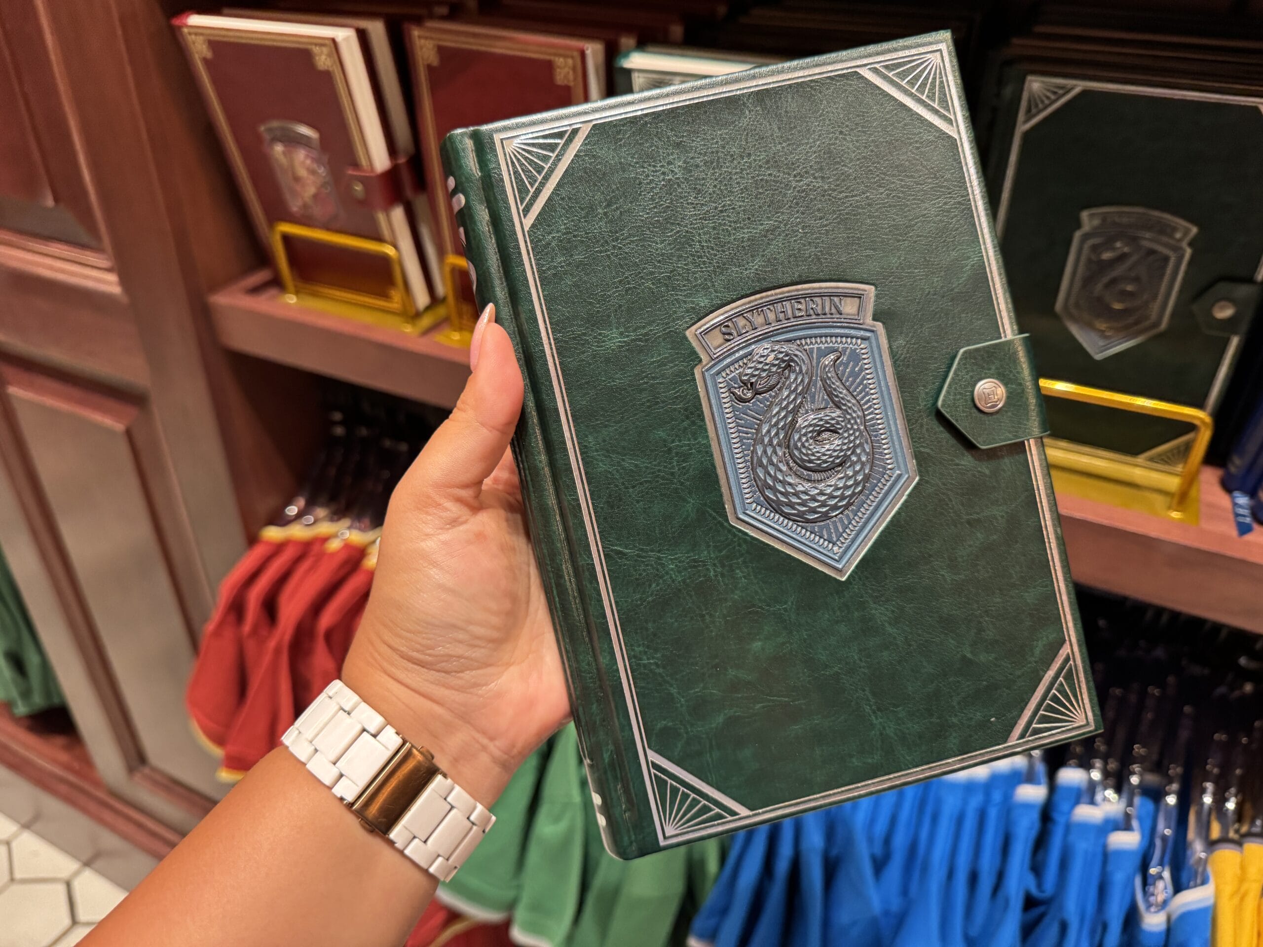 A hand holds a green Slytherin notebook, part of Wizarding World of Harry Potter merchandise, in a store display.
