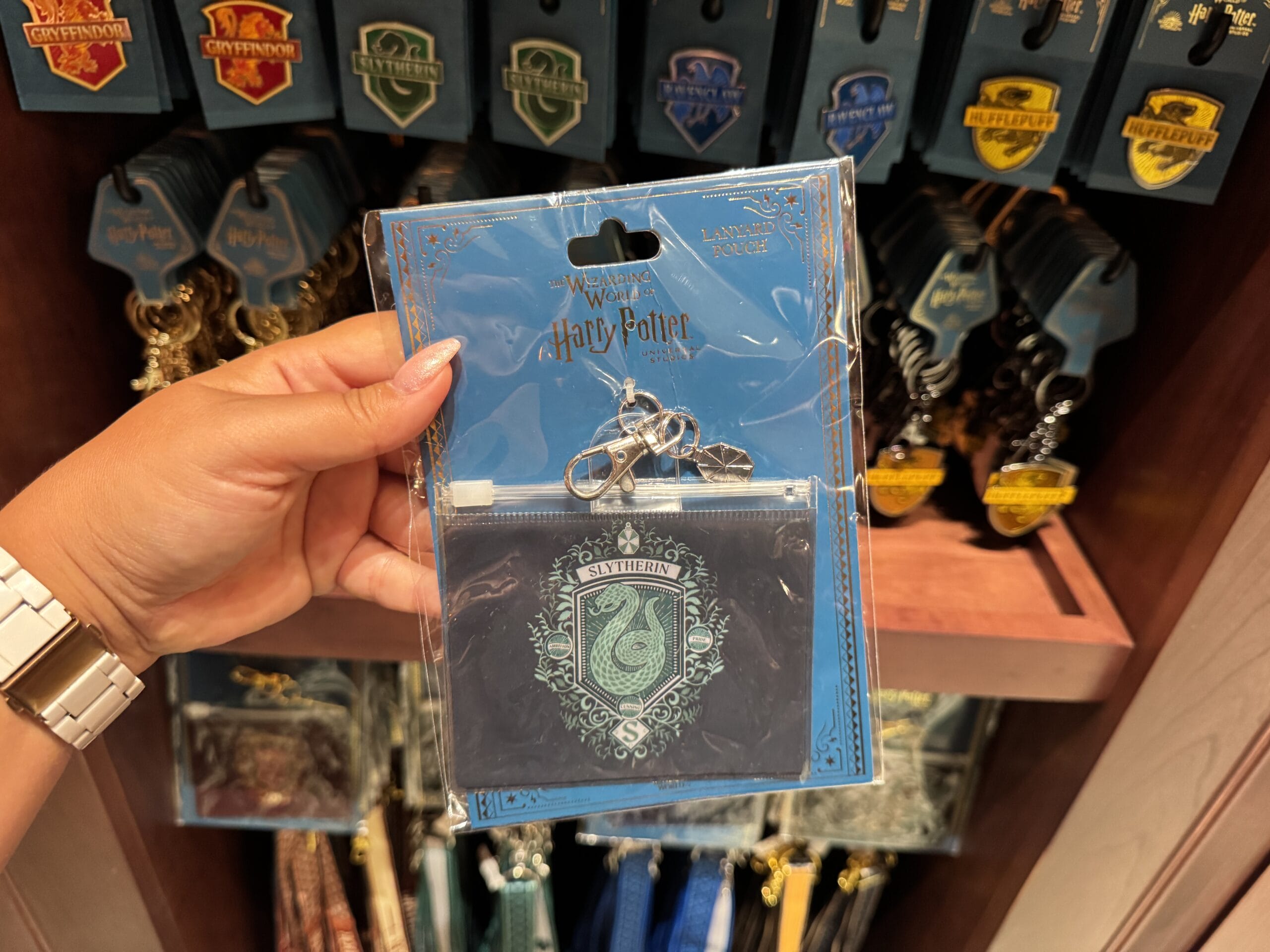 A hand holds Wizarding World of Harry Potter merchandise—a Slytherin coin pouch and keychain—at a souvenir shop display.