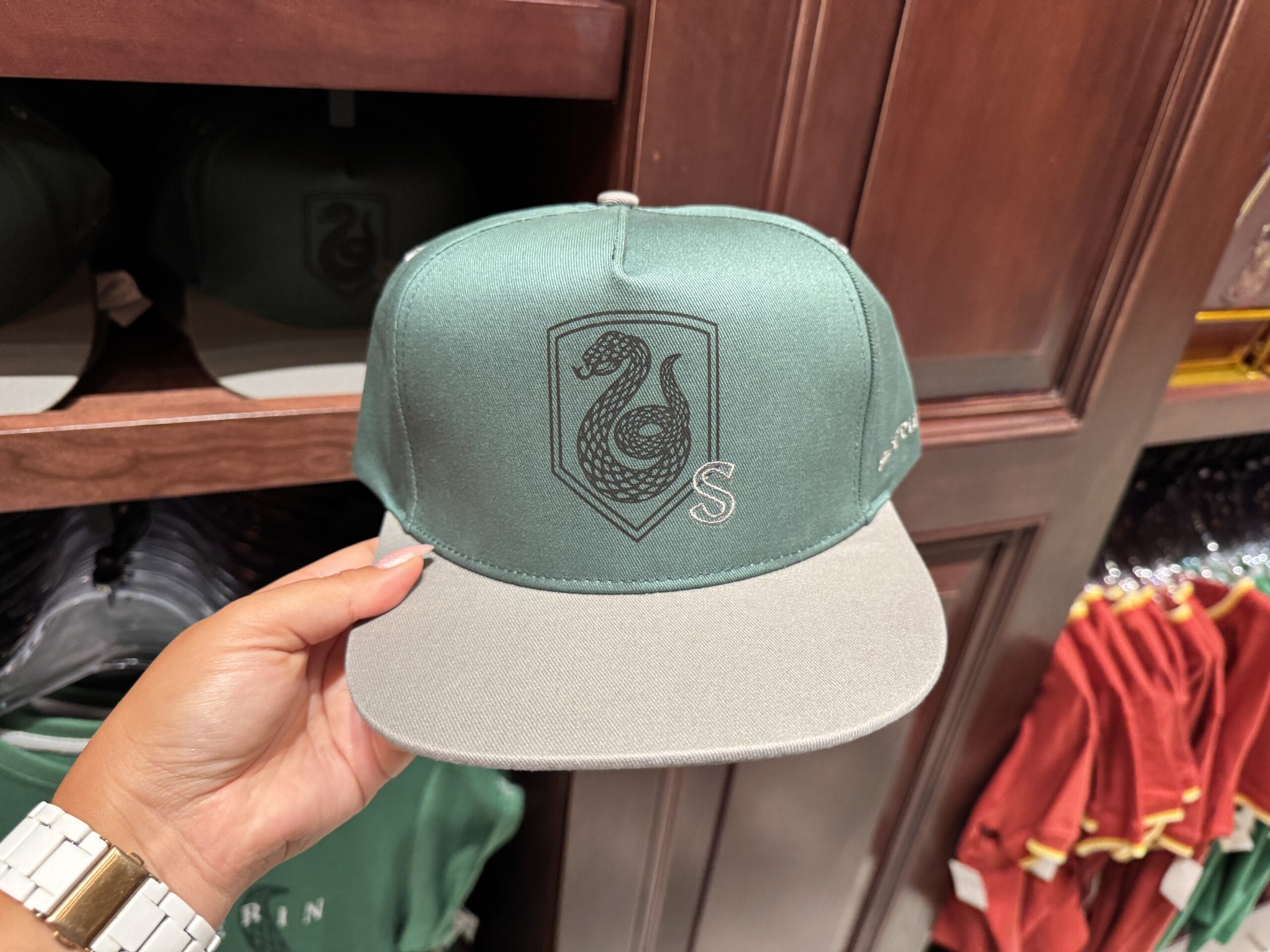 A hand holds Wizarding World of Harry Potter merchandise—a green cap with a black serpent "S" emblem, displayed in a store.