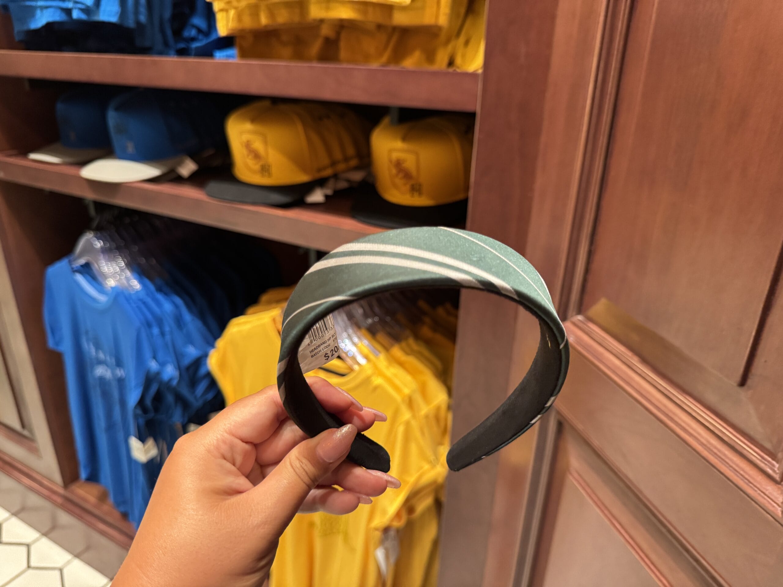 A hand holds a green and white striped headband amid Epic Universe shelves with Wizarding World of Harry Potter merchandise.