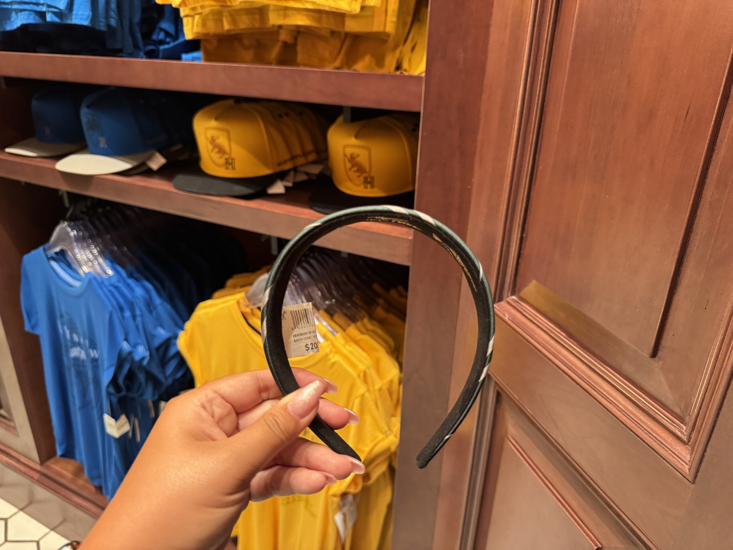 A hand holds a plain black headband in a store selling Wizarding World merchandise with yellow hats and blue shirts behind.
