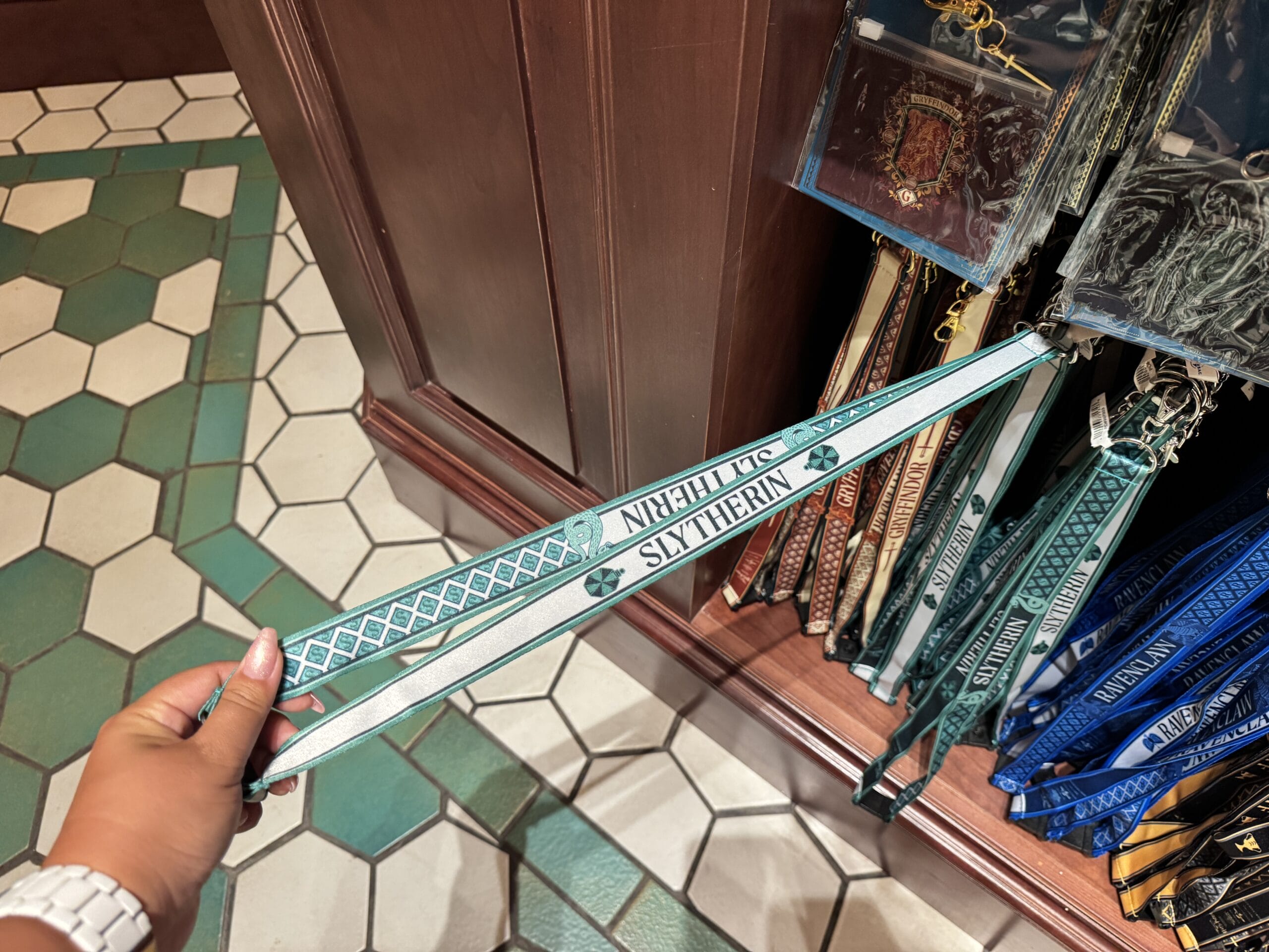 A hand holds a green Slytherin lanyard in front of Epic Universe's Wizarding World of Harry Potter merchandise shelves.