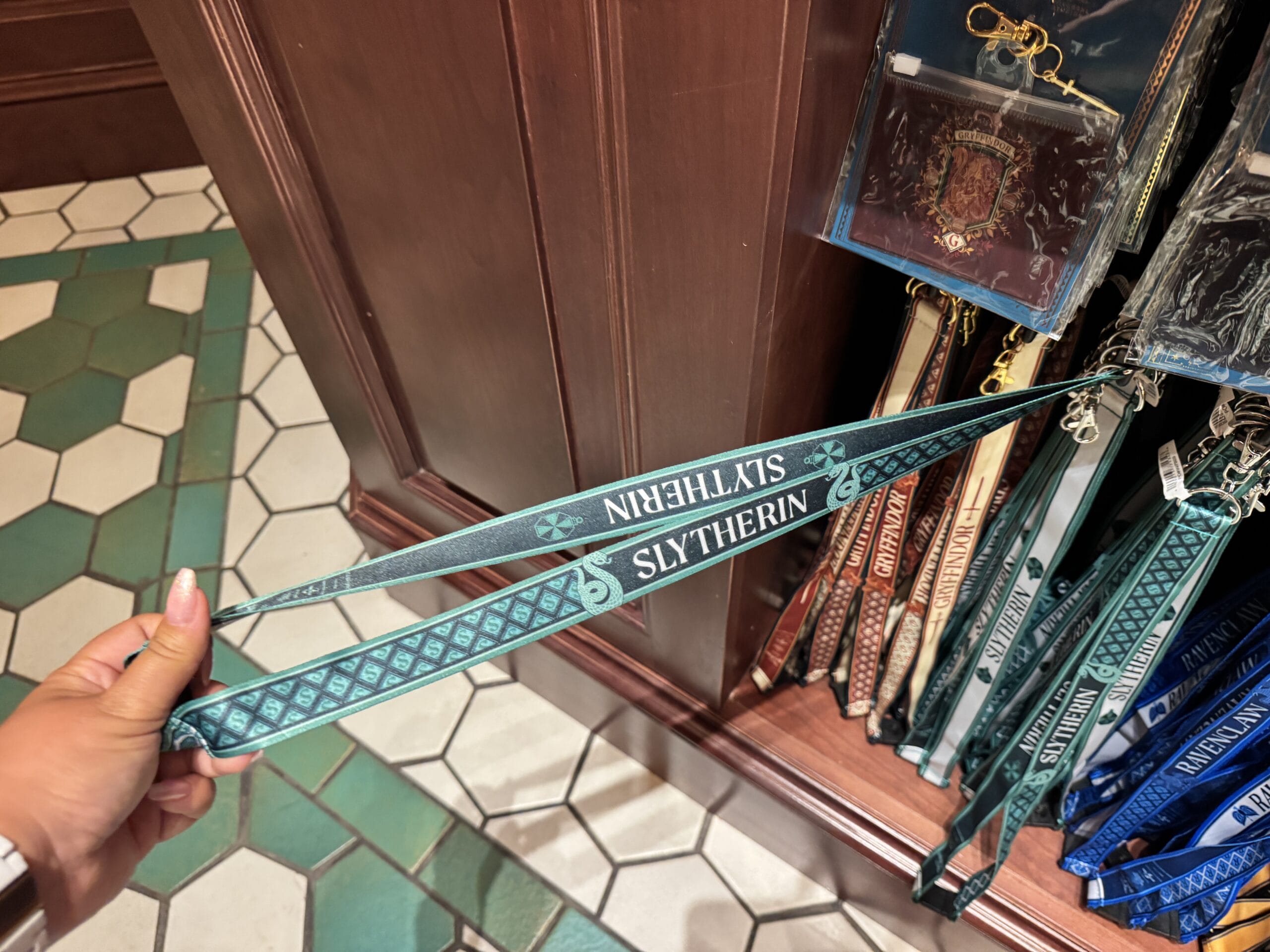 A hand holds a green "Slytherin" lanyard among Wizarding World of Harry Potter merchandise in a tiled-floor store.