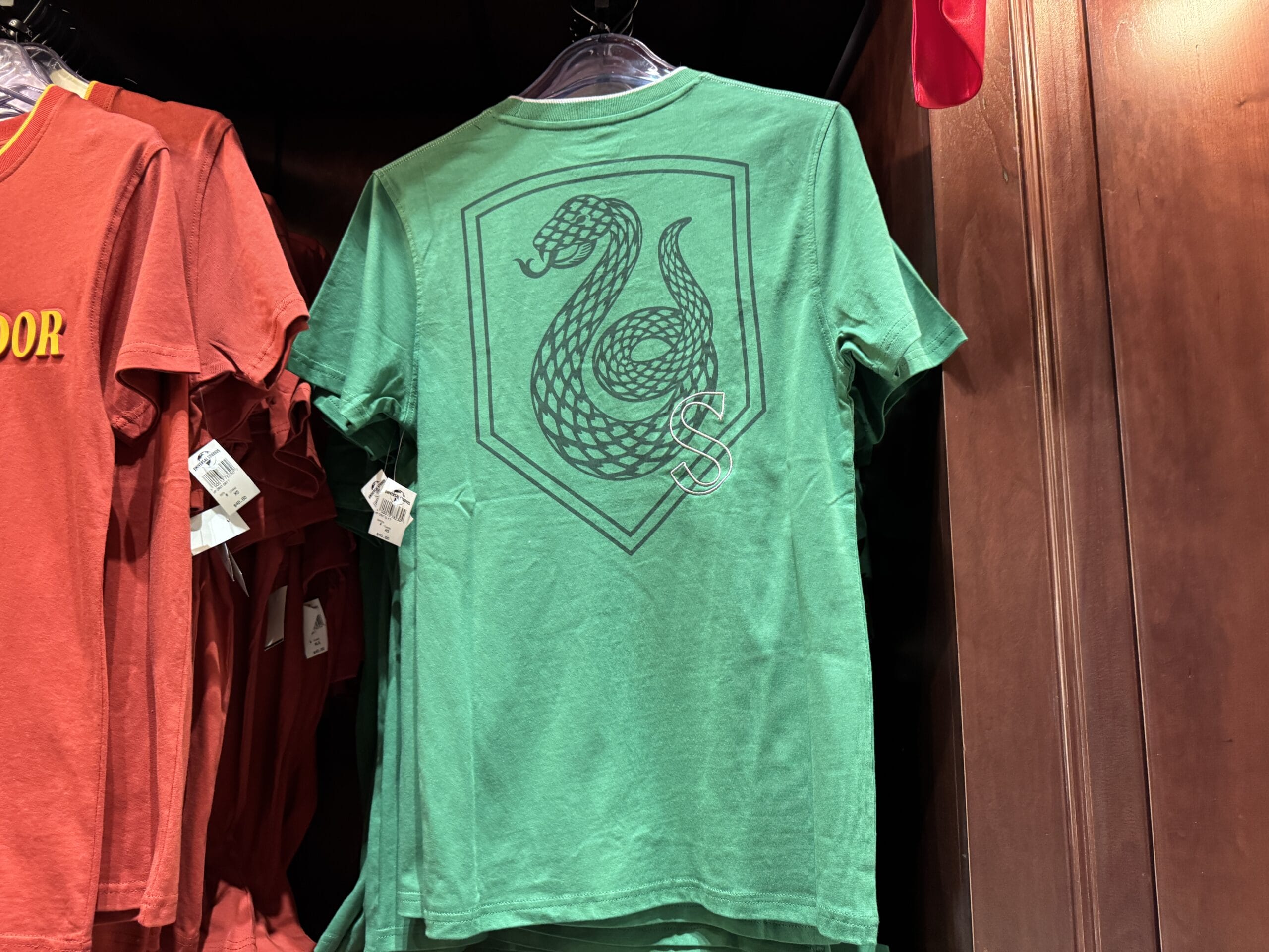 Green T-shirt with a black serpent emblem, part of Epic Universe’s Harry Potter merchandise, displayed on a rack.
