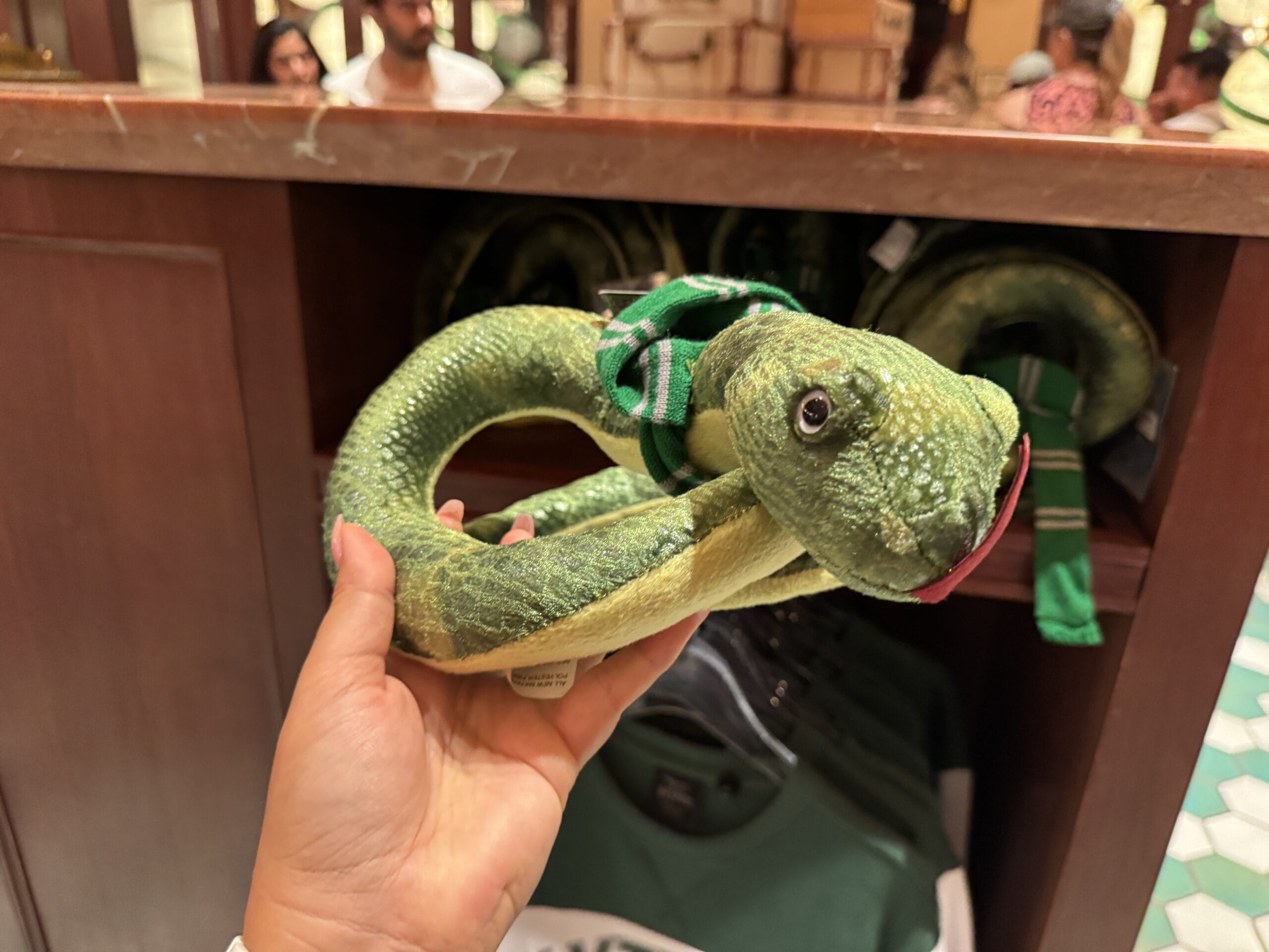 A hand holds a green plush snake with a green and silver scarf—perfect Harry Potter merchandise for Epic Universe fans.