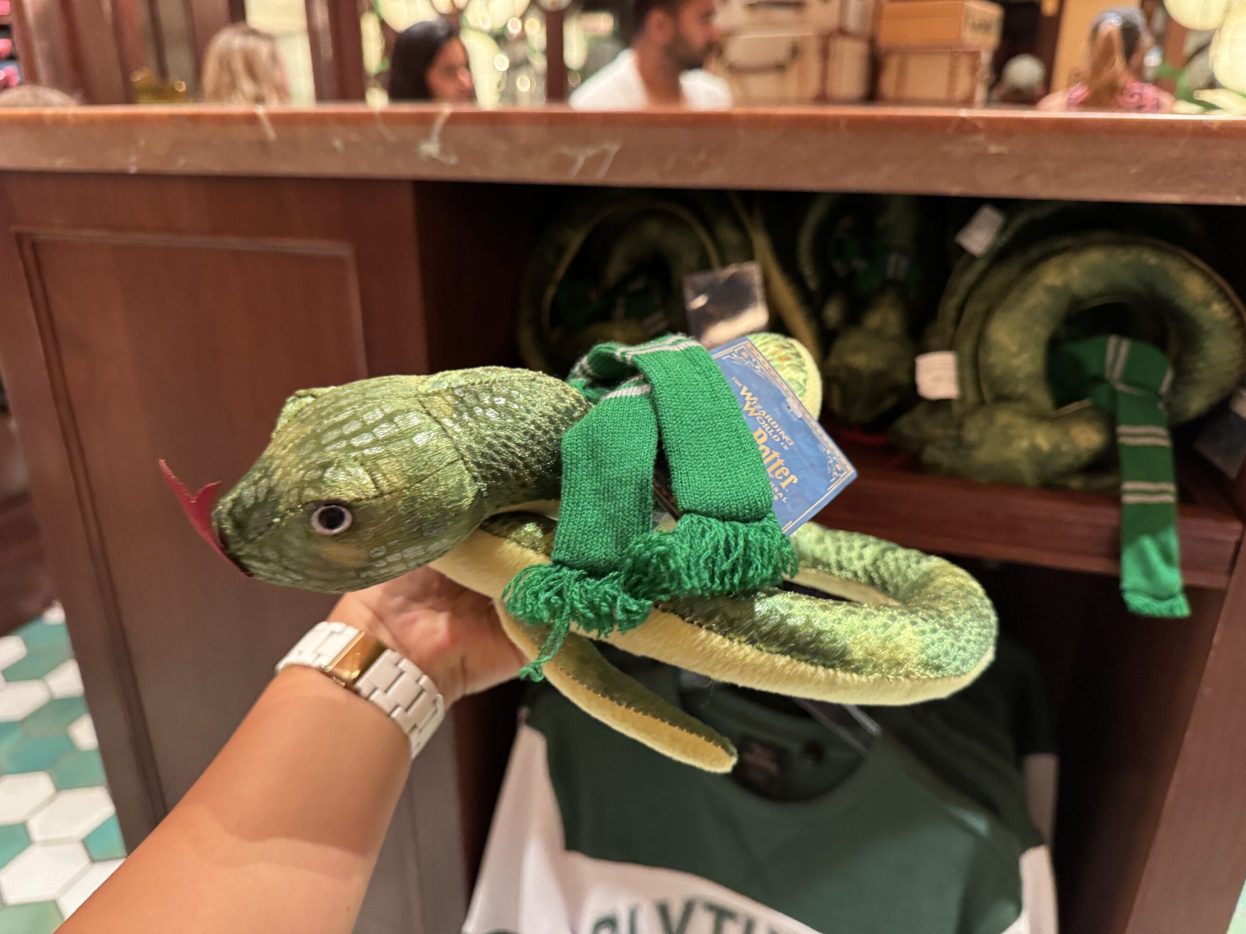 A hand holding a green snake plush with scarf, against shelves of Wizarding World of Harry Potter merchandise.
