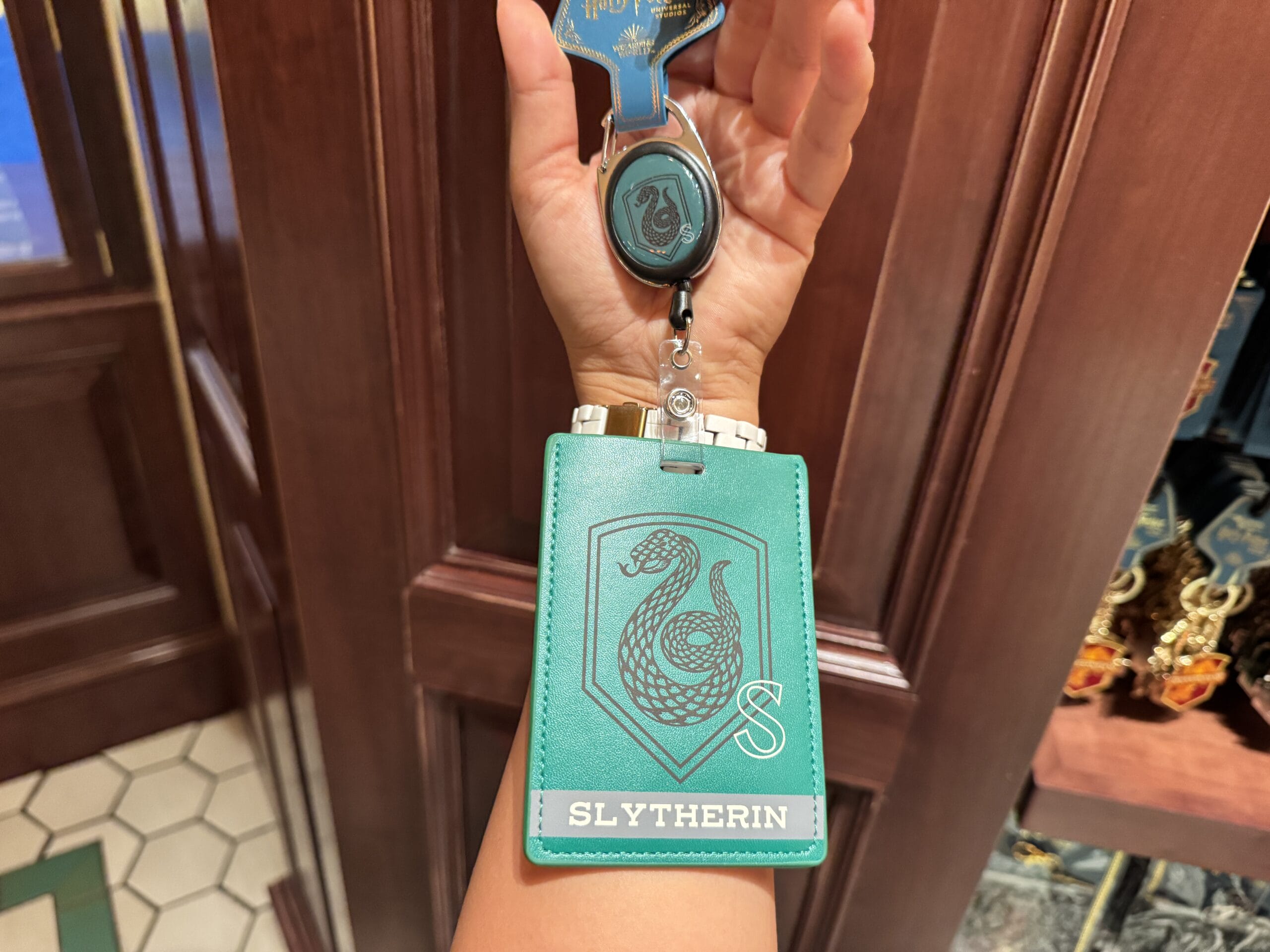 A hand holds a Slytherin badge holder—authentic Harry Potter merchandise—in front of a wooden wall.