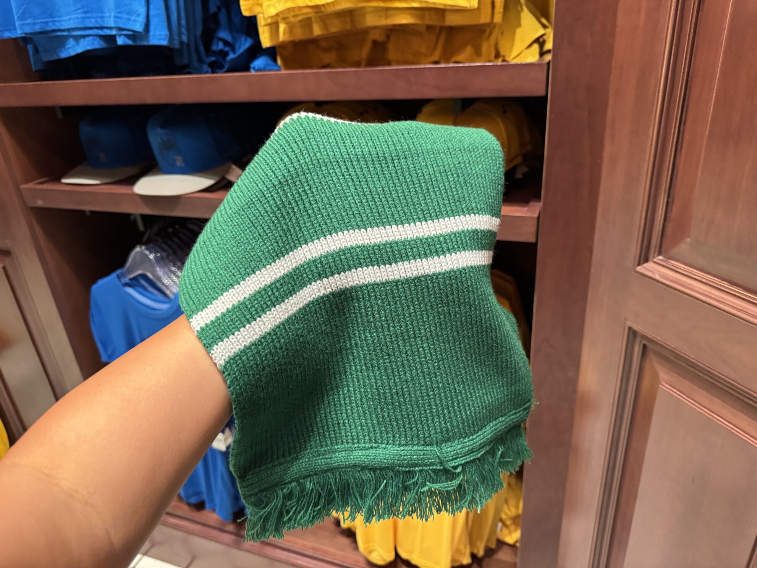 A hand holds a green knit scarf with white stripes—classic Wizarding World Harry Potter merchandise—in a store.