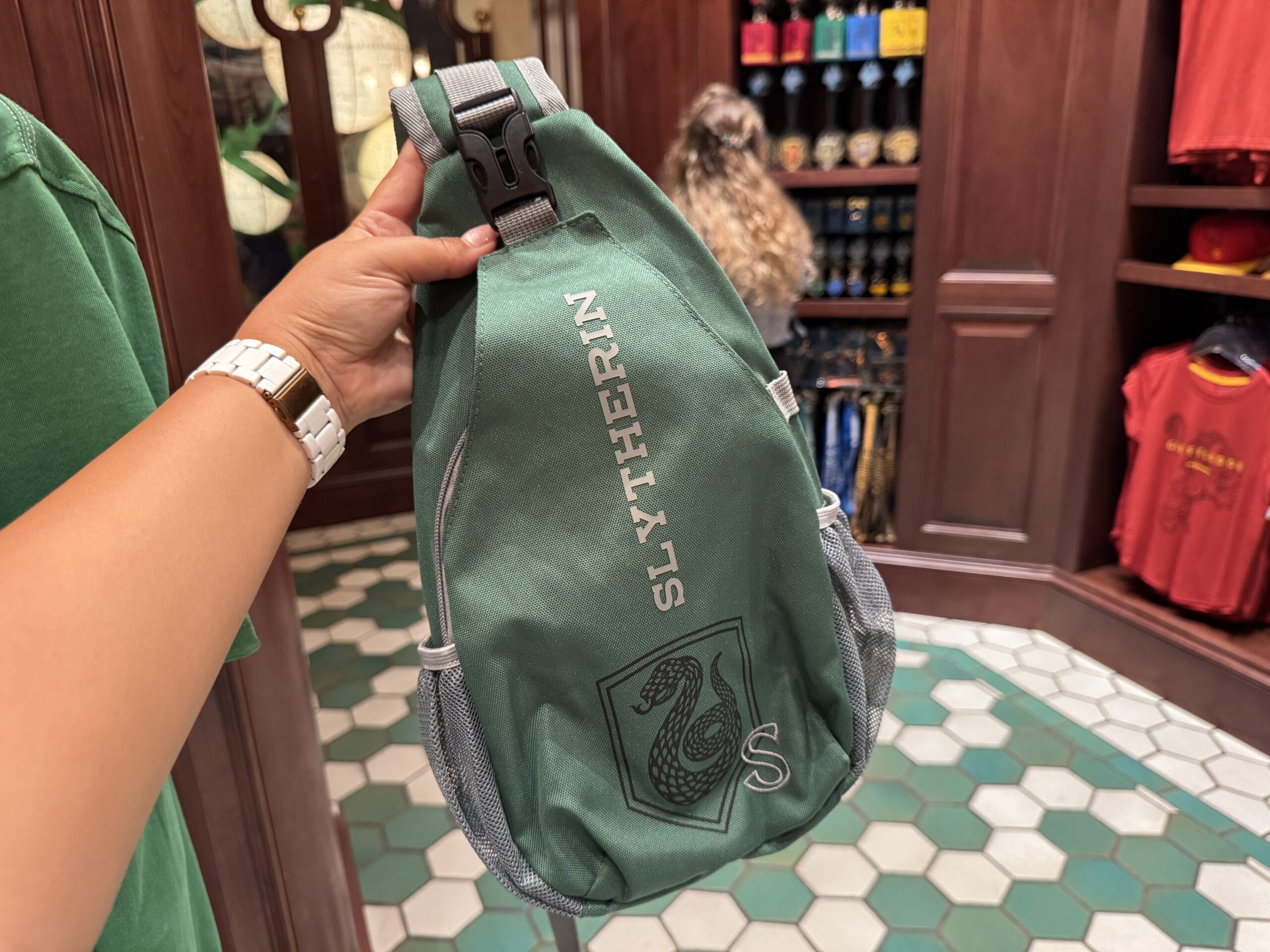 A hand holds a green Slytherin sling bag with a snake emblem—perfect Wizarding World of Harry Potter merchandise.