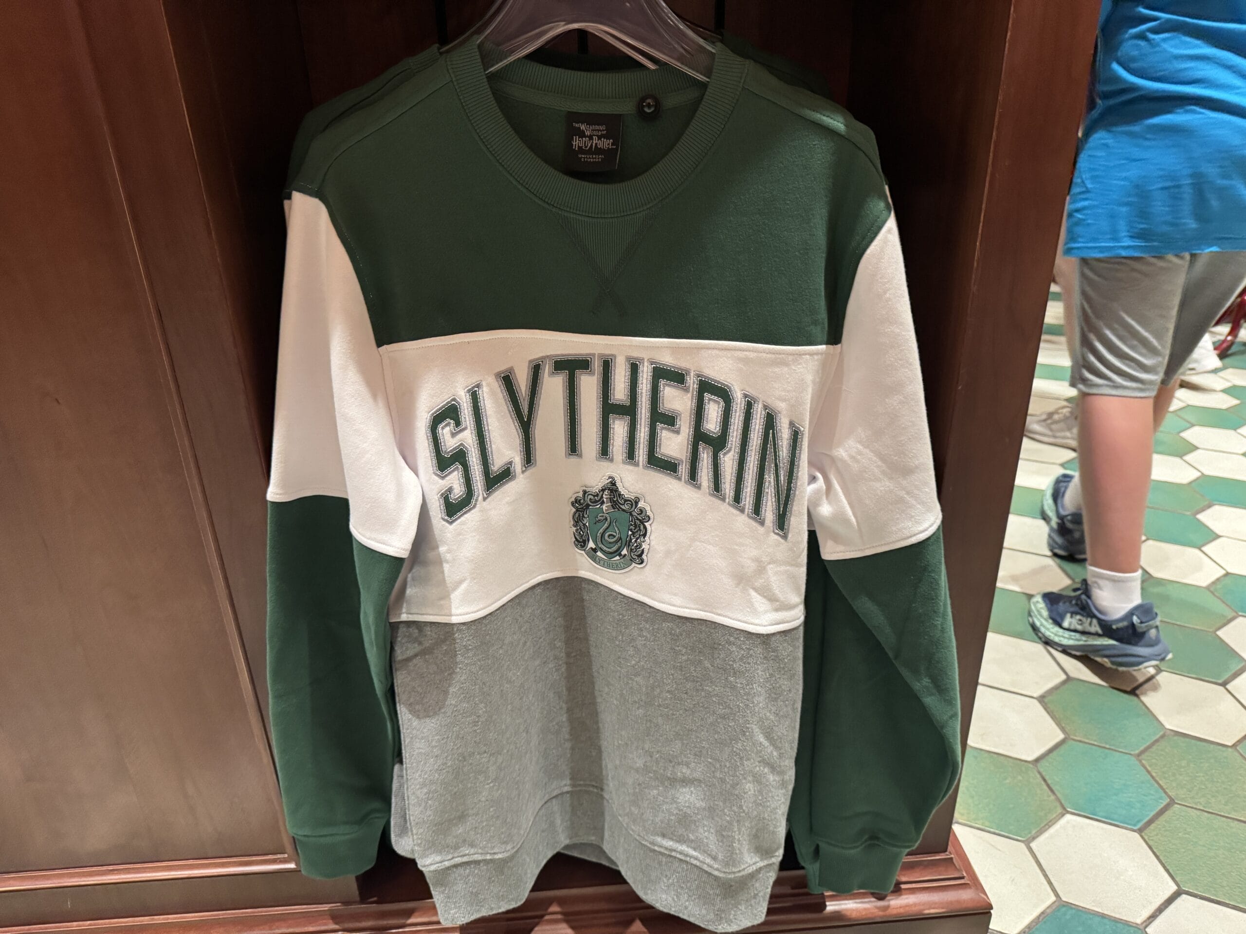 A green, white, and gray Slytherin sweatshirt from the Wizarding World of Harry Potter merchandise hangs on a store rack.
