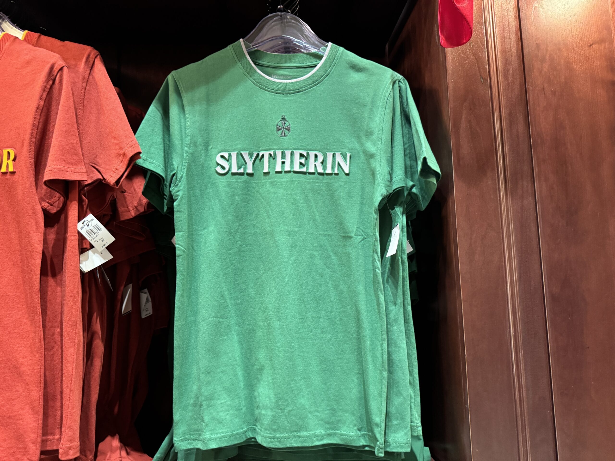 Green T-shirt with "SLYTHERIN" print, classic Wizarding World of Harry Potter merchandise, hanging beside a red shirt.