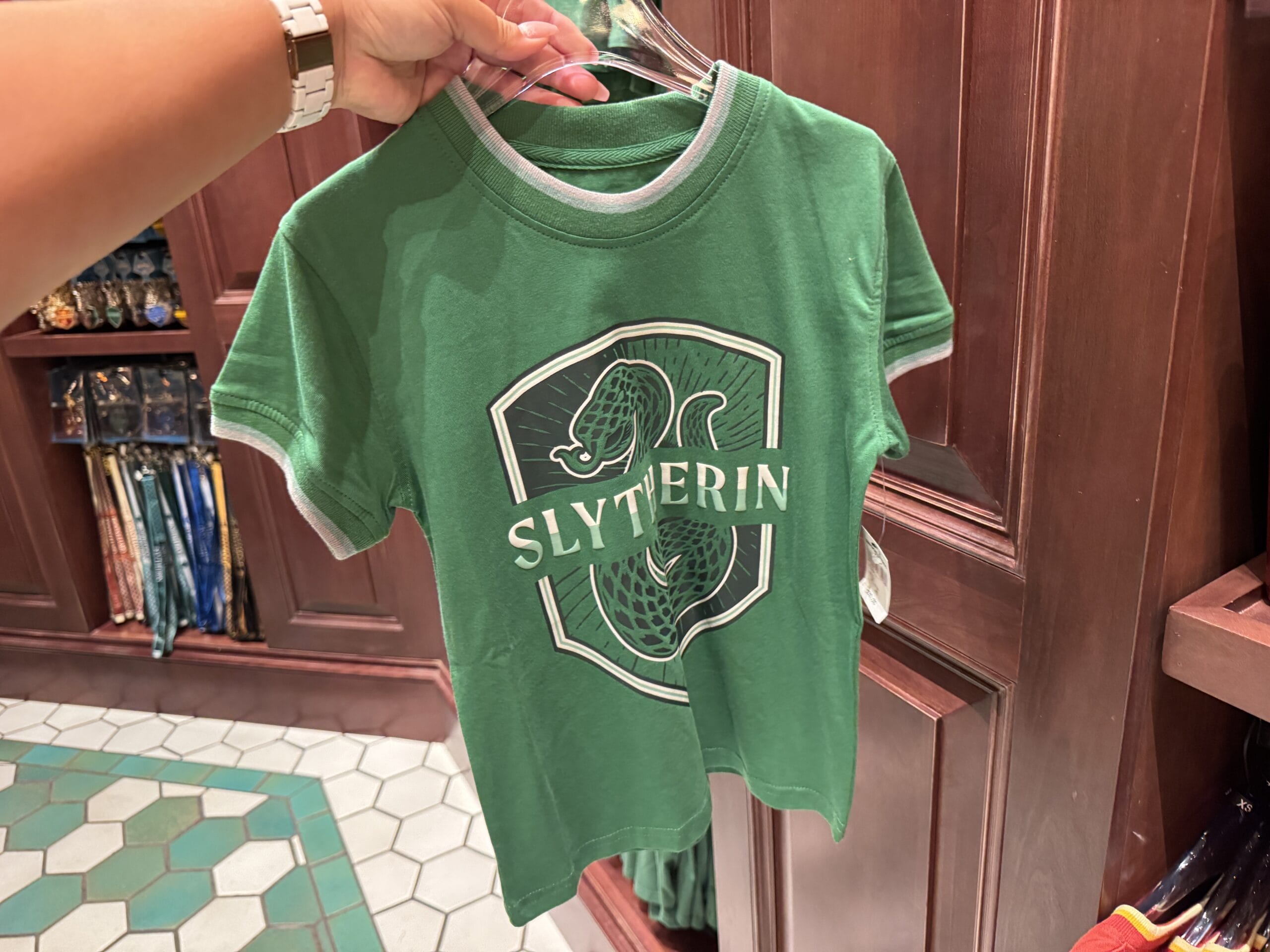 A green Slytherin t-shirt with a snake emblem, part of exclusive Harry Potter merchandise, is held up in a store.