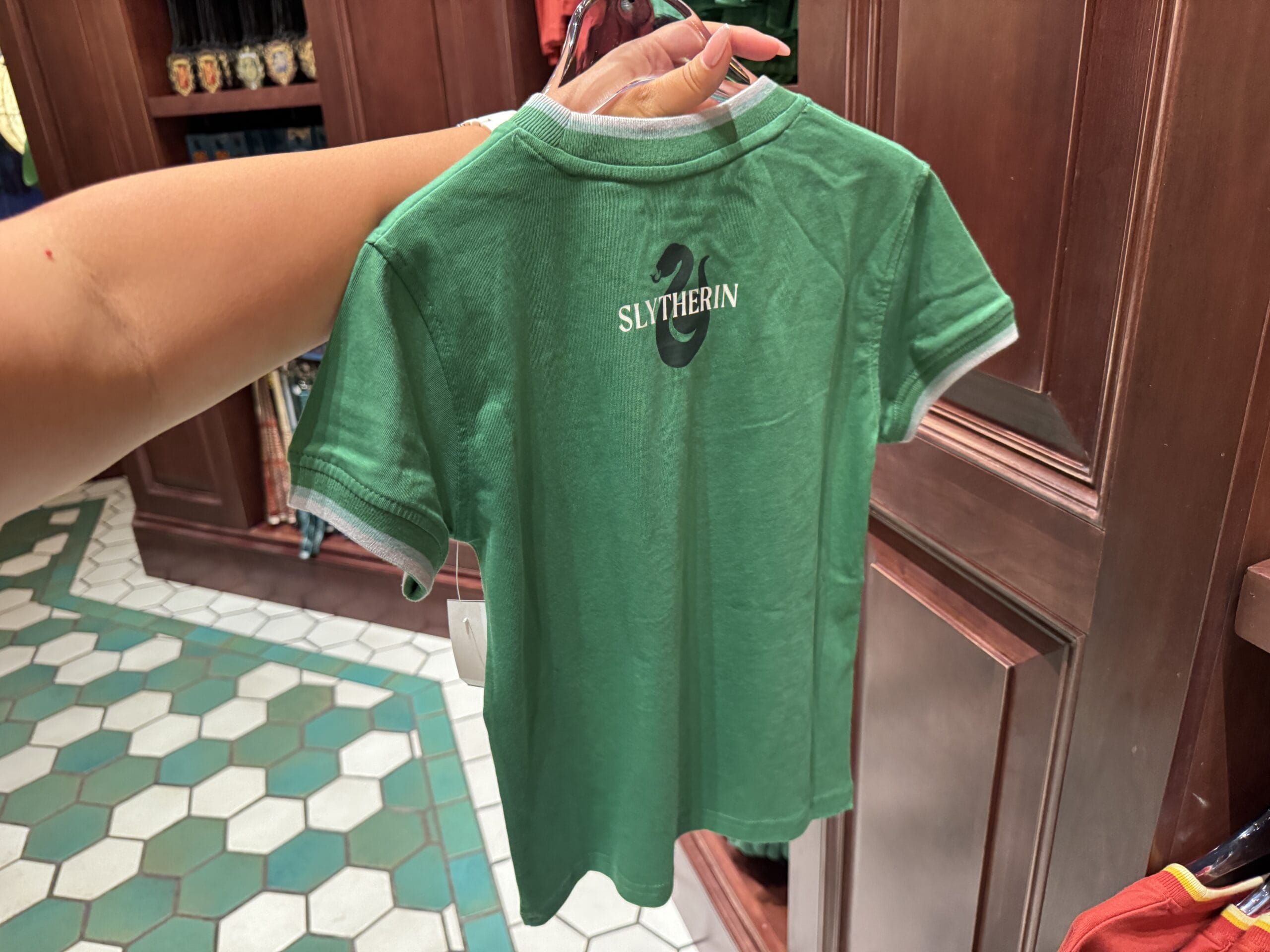 A hand holds a green children's T-shirt with "SLYTHERIN" on the back—Harry Potter merchandise found in-store.