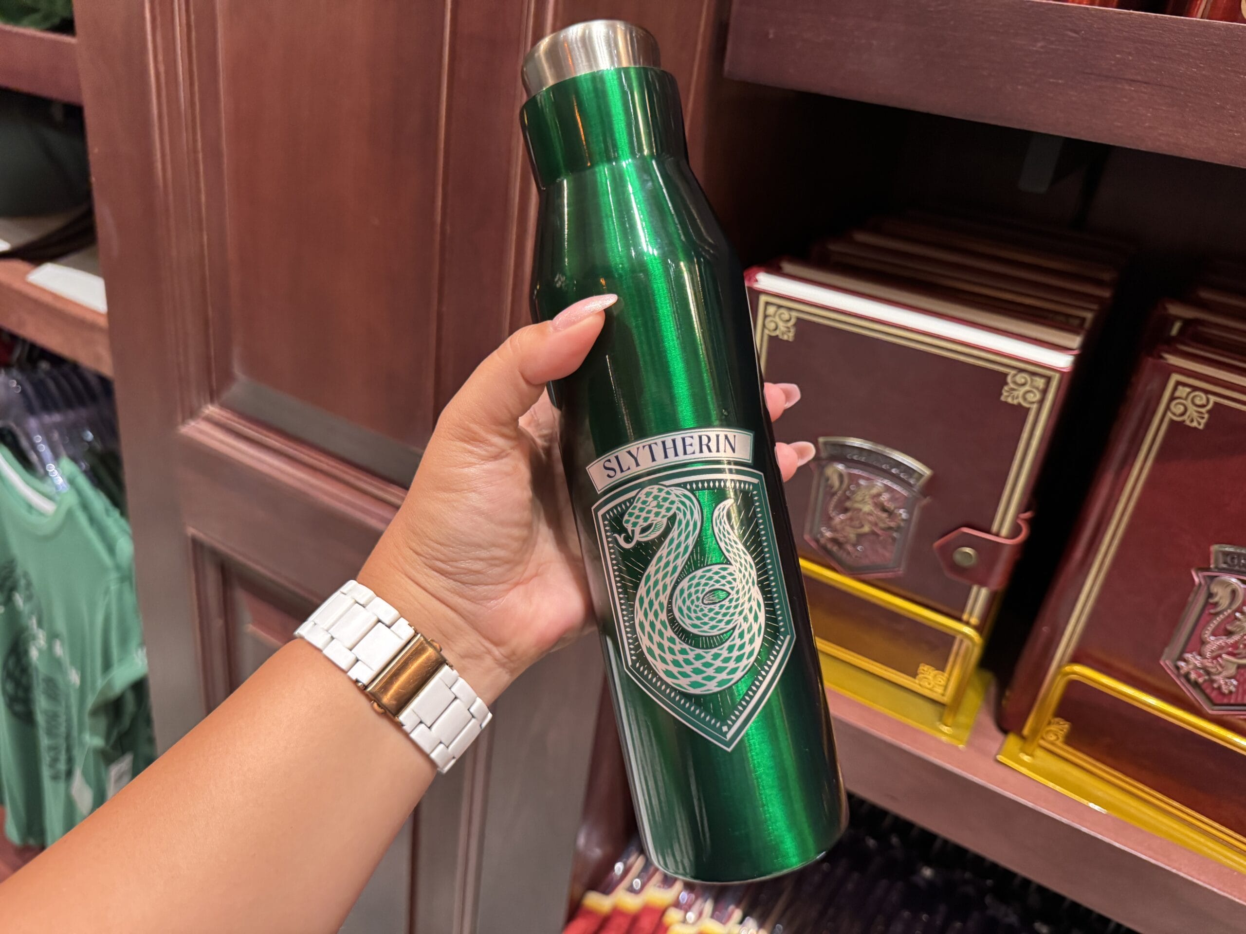 A hand holds a green water bottle with a silver Slytherin emblem, exclusive Wizarding World Harry Potter merchandise.
