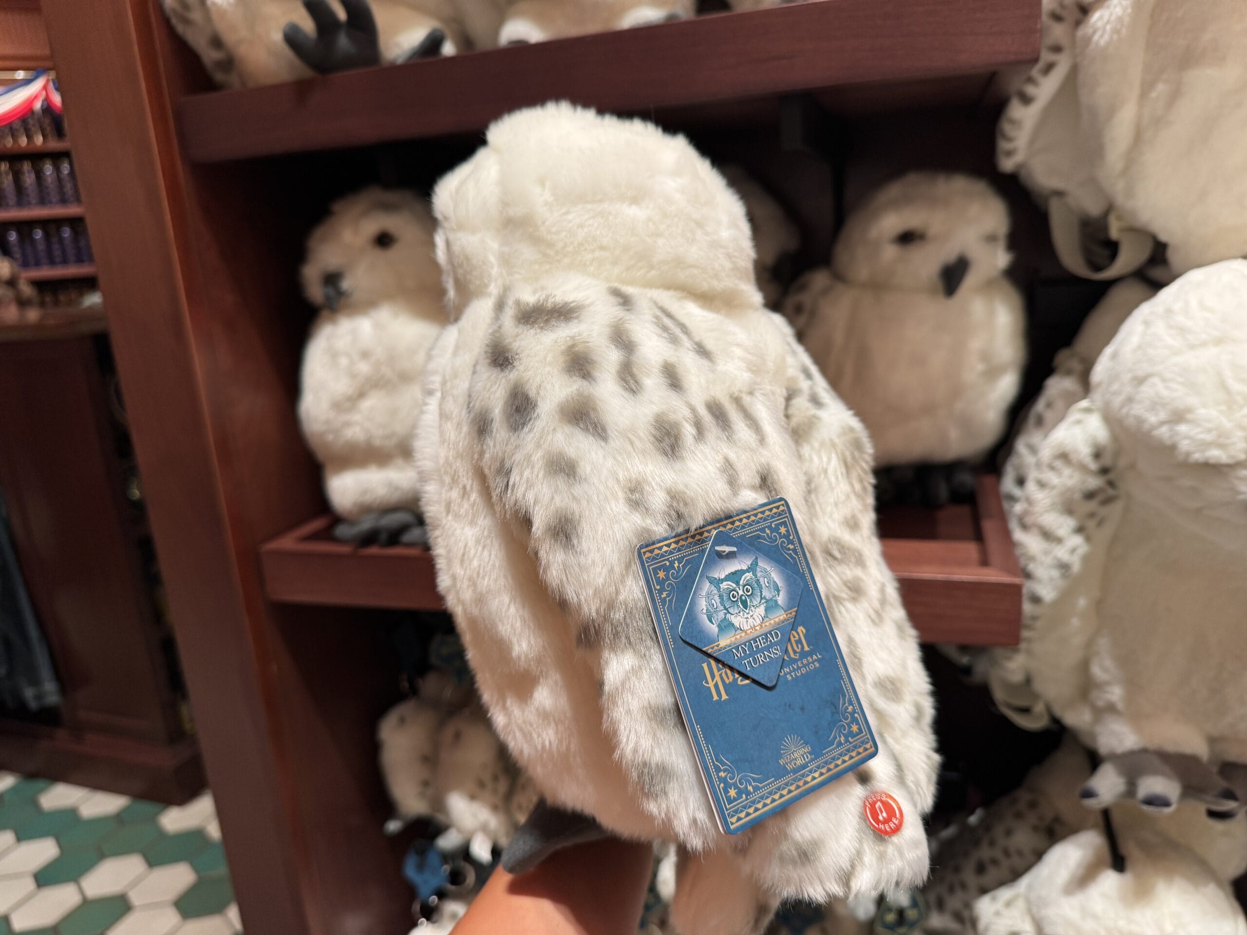 A hand holds a plush snowy owl toy with a blue Hogwarts tag—magical Harry Potter merchandise among others on the shelf.