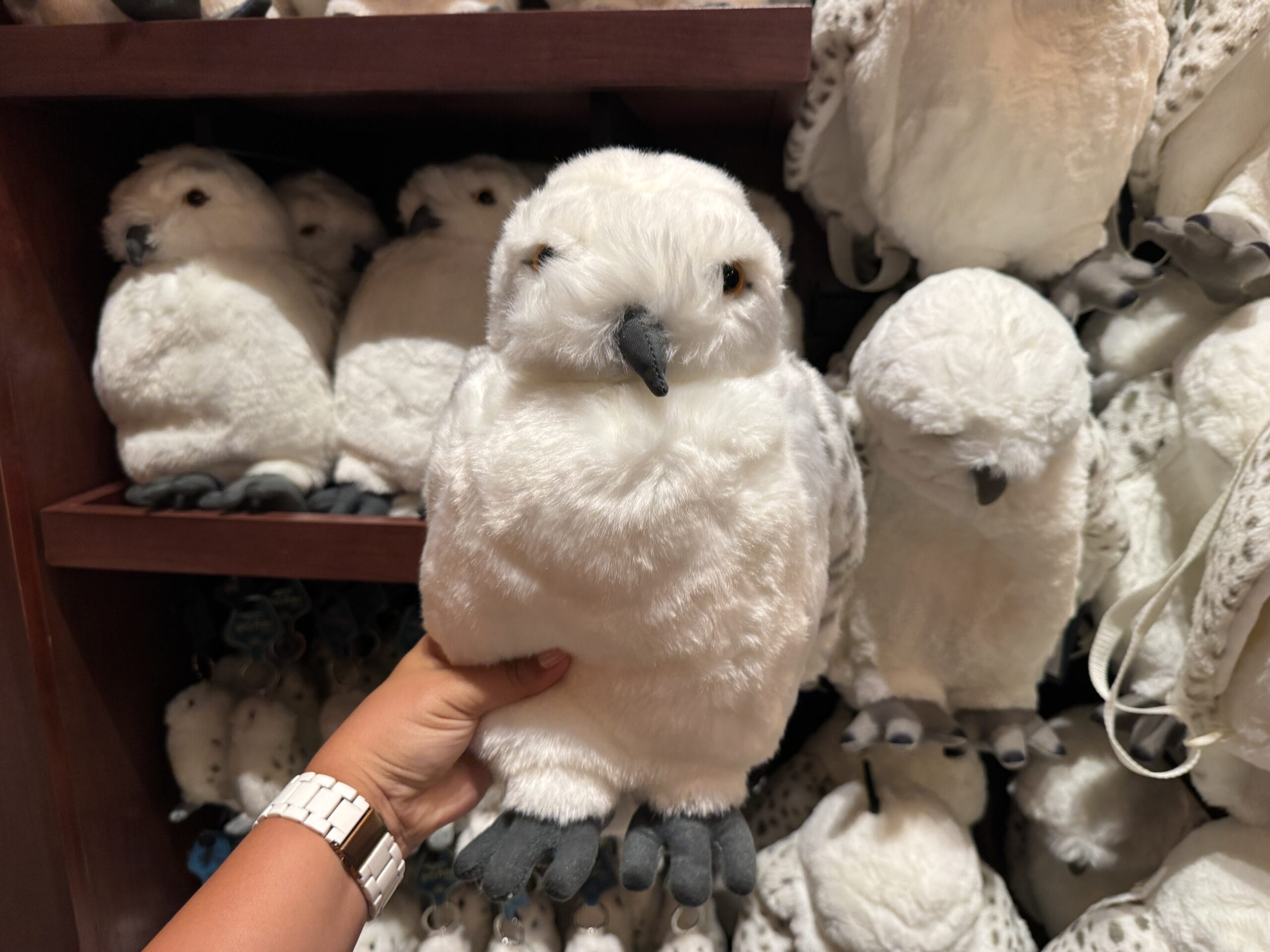 A hand holds a plush snowy owl toy, part of Tour En Floo merchandise, in front of a shelf of similar stuffed owls.