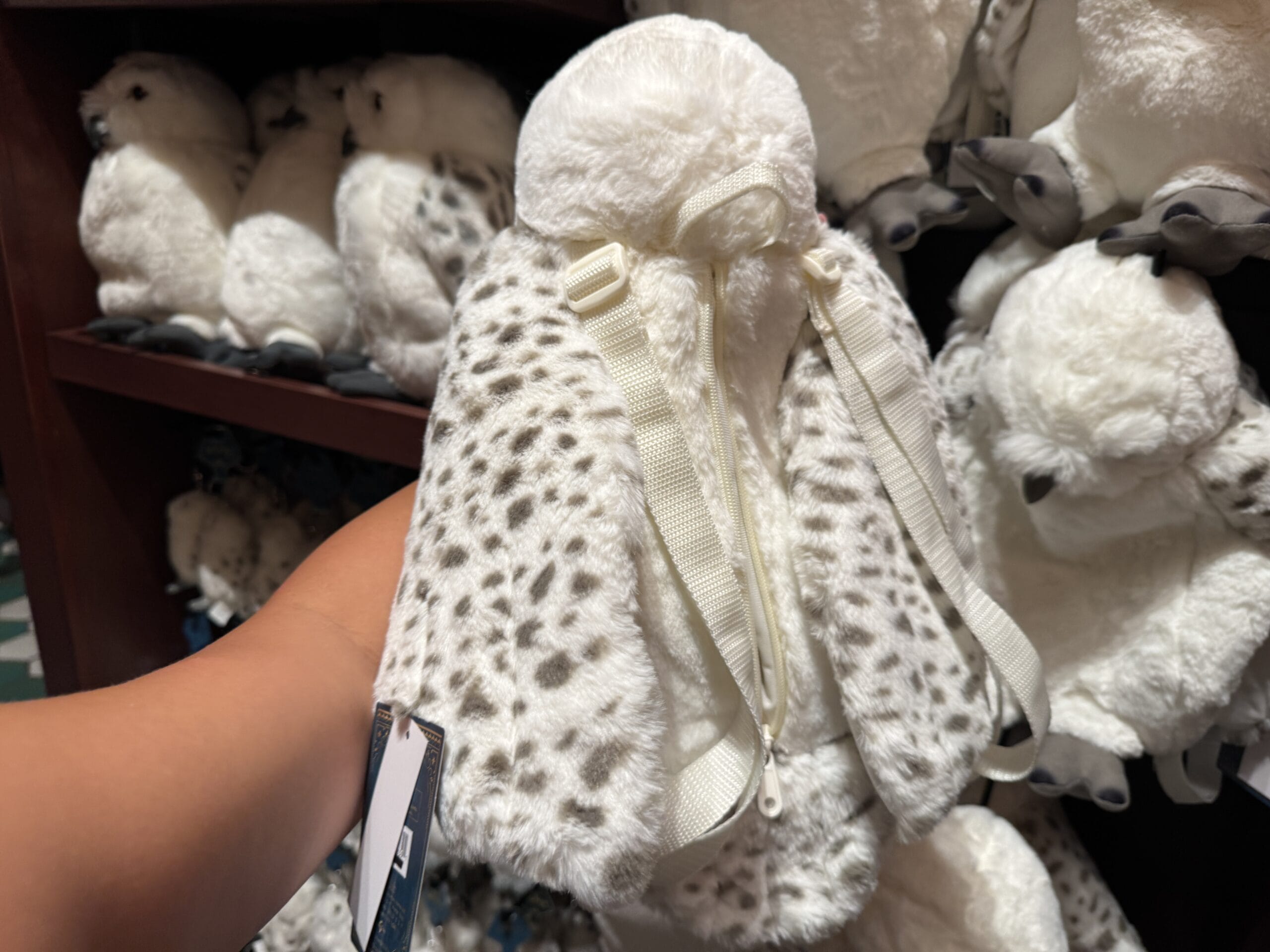 A hand holds a plush snowy owl backpack with black spots, displayed among Harry Potter merchandise on shelves.