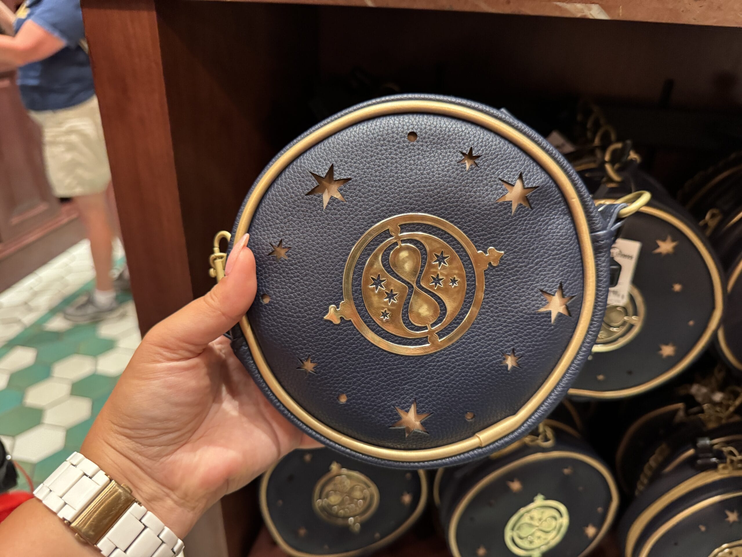 A hand holds a round, navy blue purse inspired by Epic Universe, adorned with gold stars and a celestial design.
