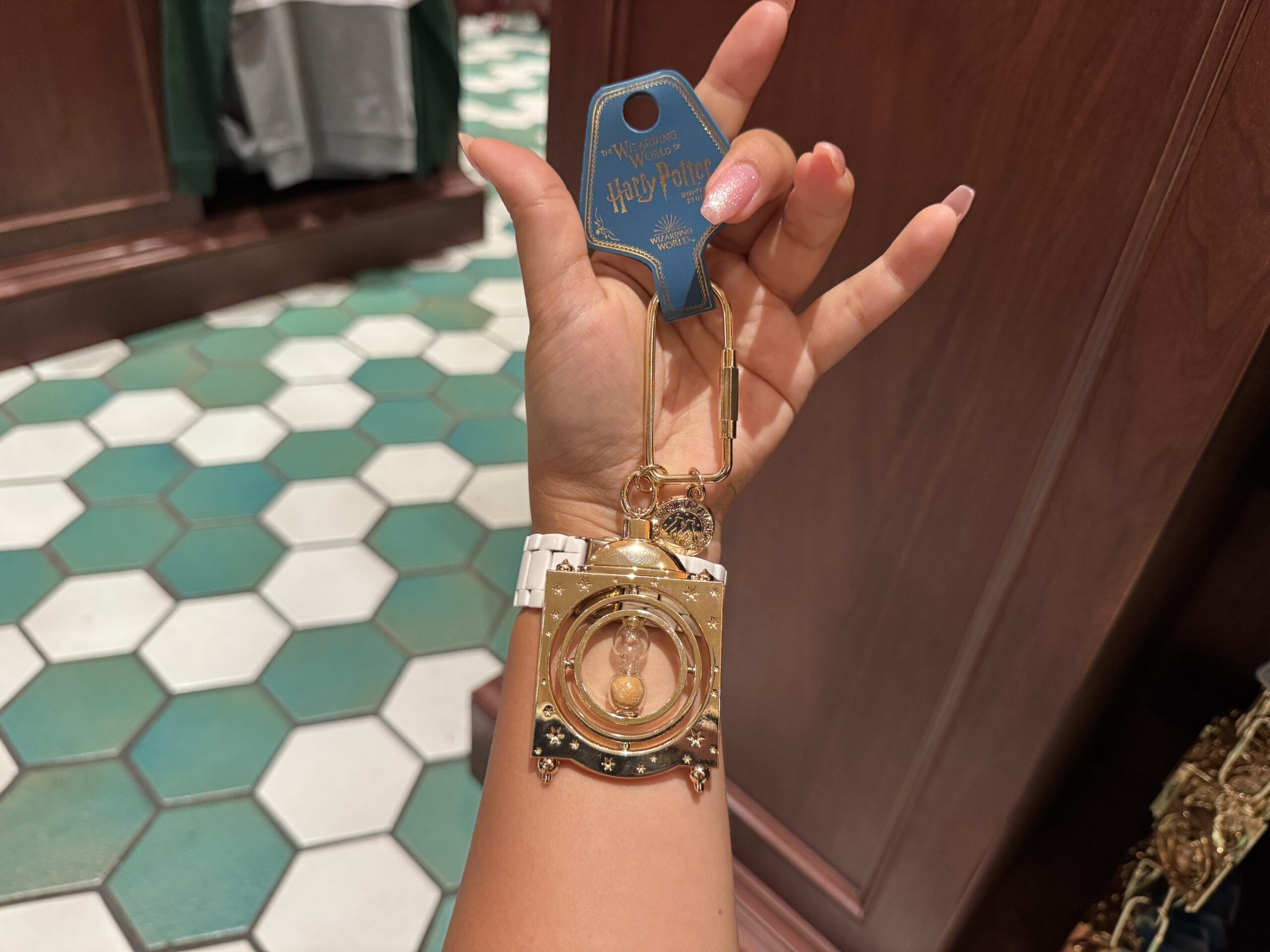 A hand holds Wizarding World Harry Potter merchandise—a gold Time-Turner keychain—against a tiled floor and wooden wall.