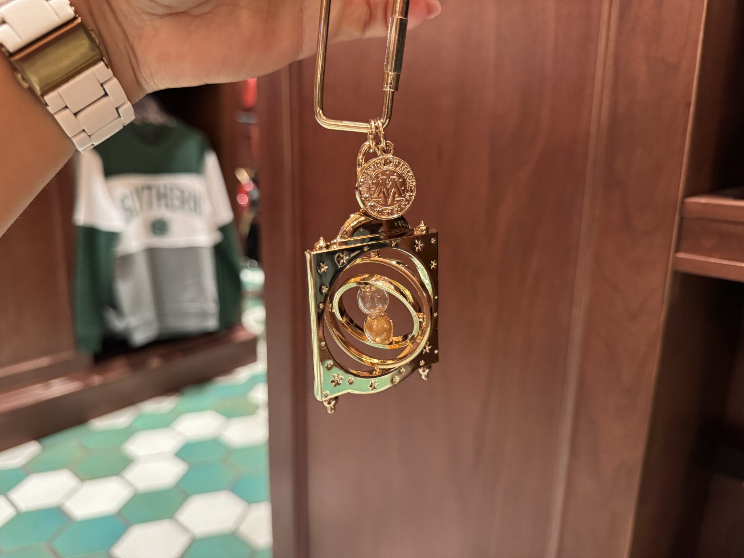 A hand holds a golden Wizarding World time-turner keychain in a store with wooden shelves and green clothing behind.
