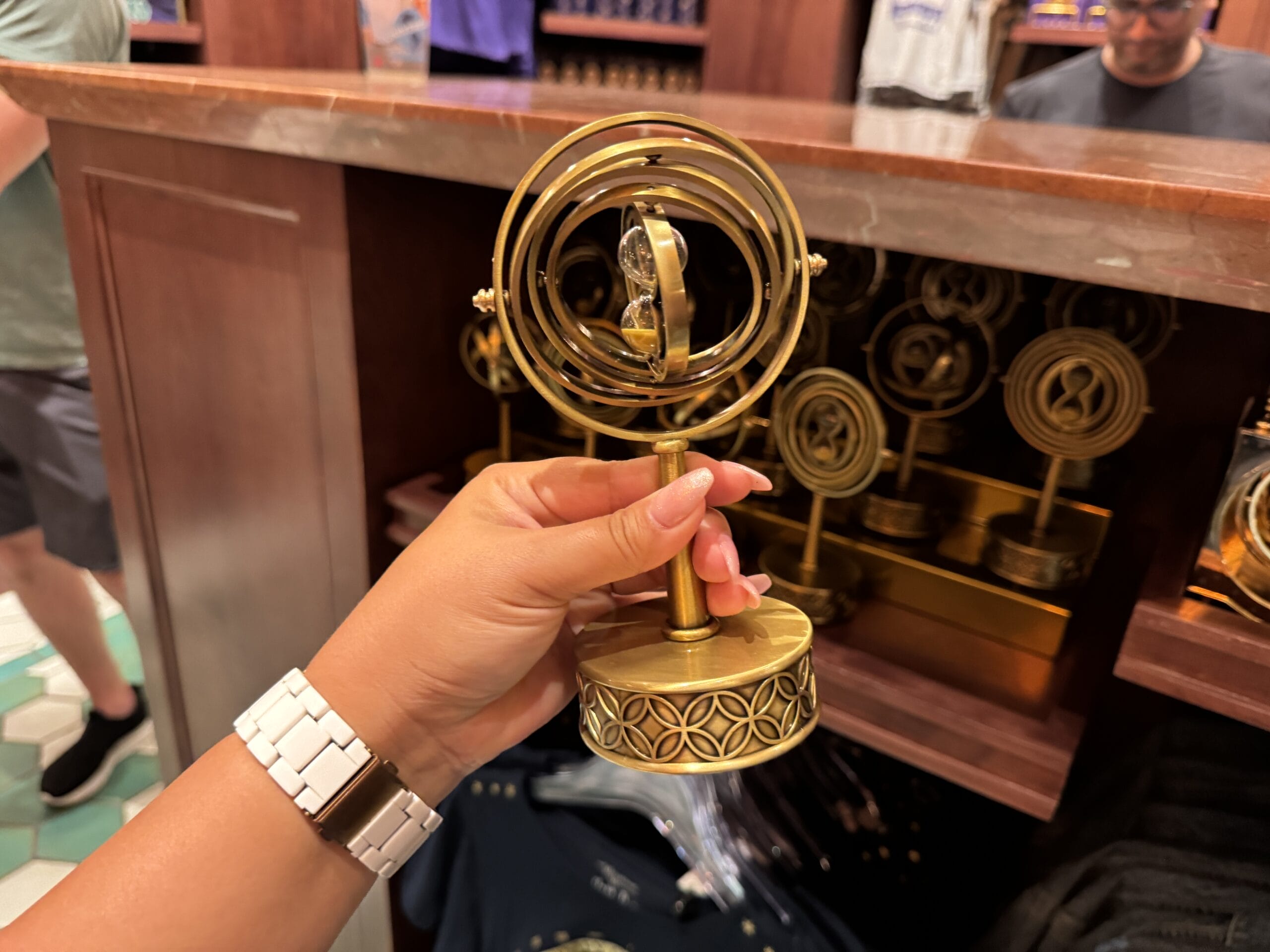 A hand holds a golden time turner replica, surrounded by Wizarding World of Harry Potter merchandise on display.
