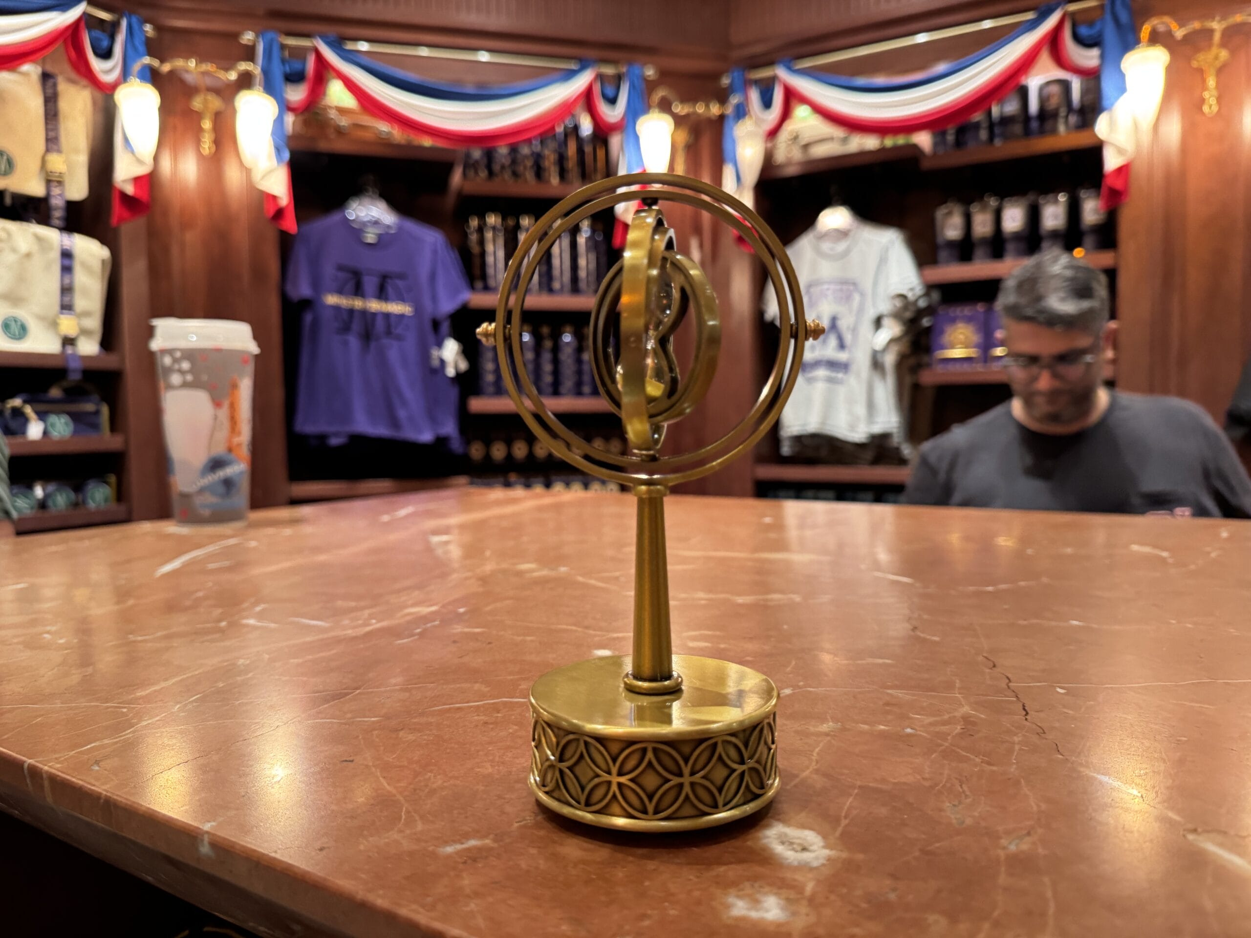 A brass gyroscope stands on a marble counter in a shop filled with shirts, mugs, and Harry Potter merchandise.