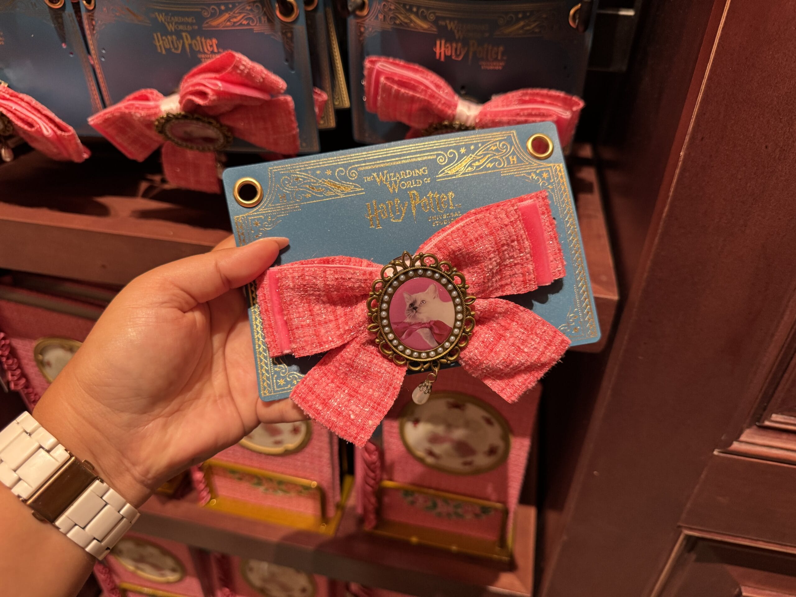 A hand holds a pink bow hair accessory with a cat photo, Wizarding World of Harry Potter merchandise, in a store display.
