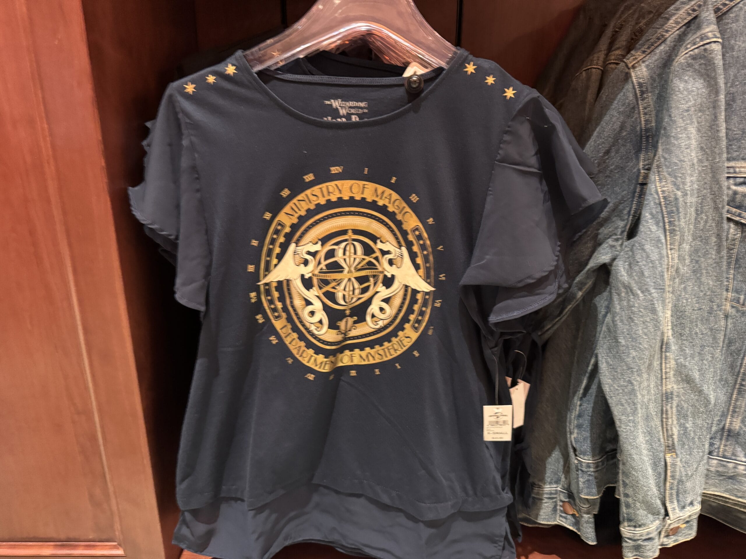 Navy blue shirt with gold "Ministry of Magic" crest, perfect Wizarding World of Harry Potter merchandise, next to a denim jacket.