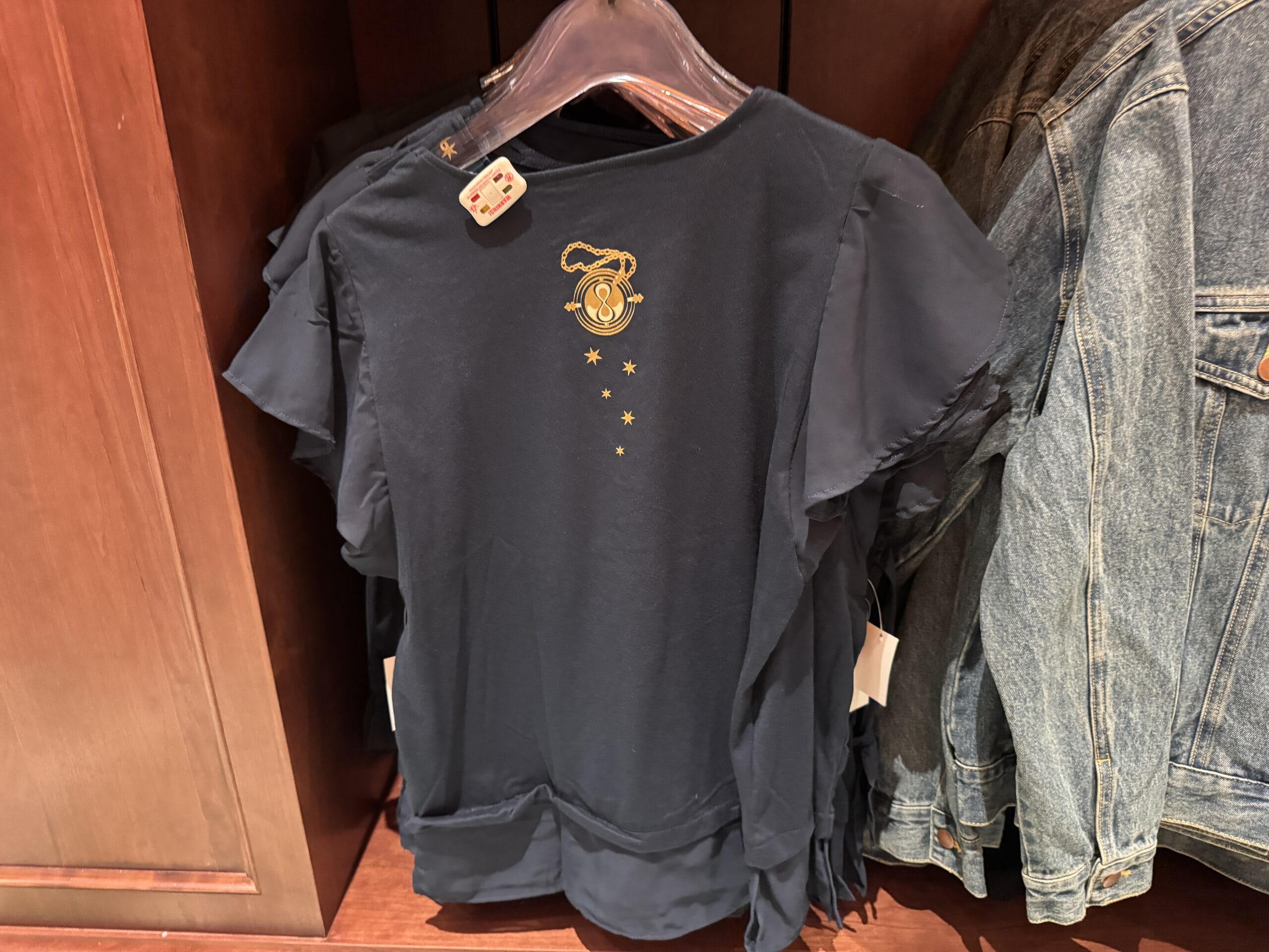 A navy blue t-shirt with a small gold serpent and stars, inspired by Wizarding World, hangs on a rack.