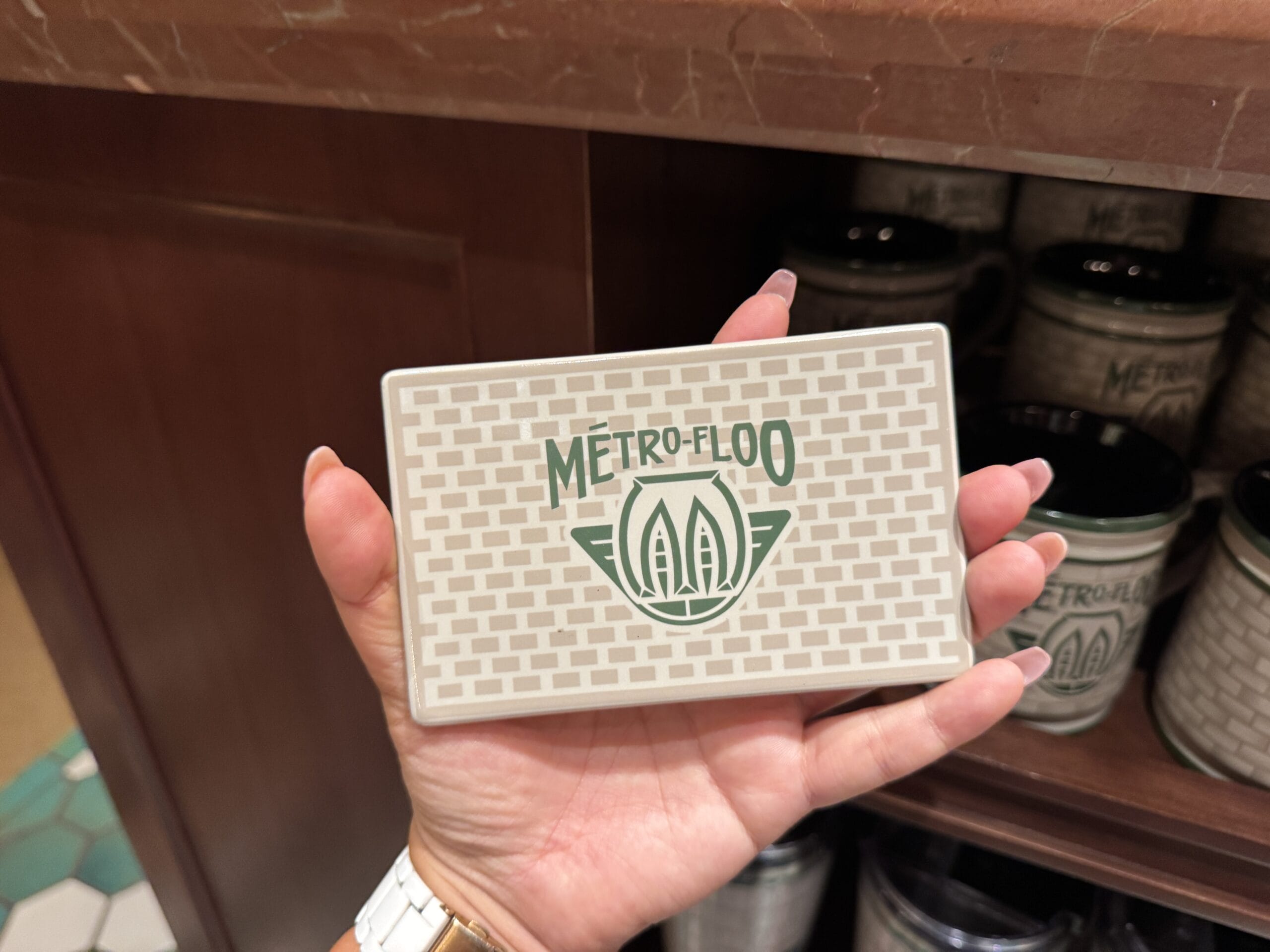 A hand holds a "METRO-FLOO" tile by matching mugs—perfect for Tour En Floo fans at Epic Universe.