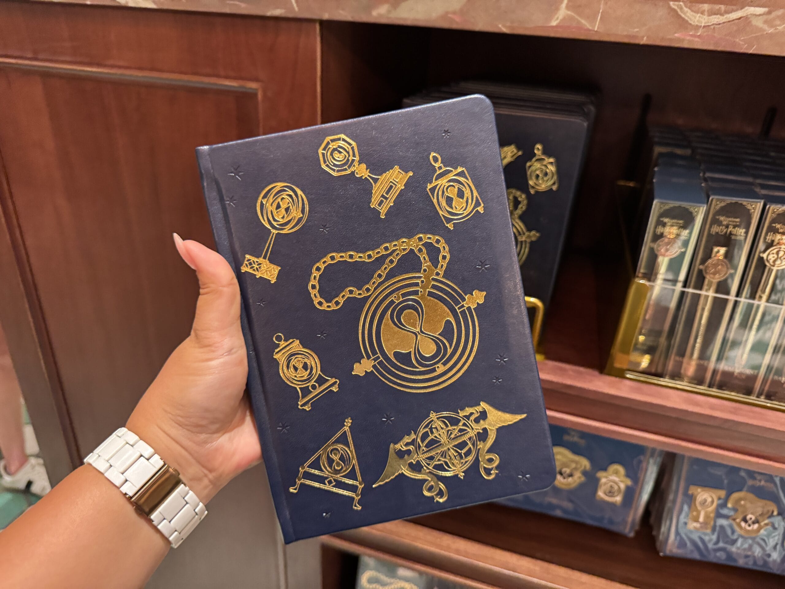 A hand holds a navy blue notebook adorned with gold magical symbols, perfect for any Wizarding World or Harry Potter merchandise fan.