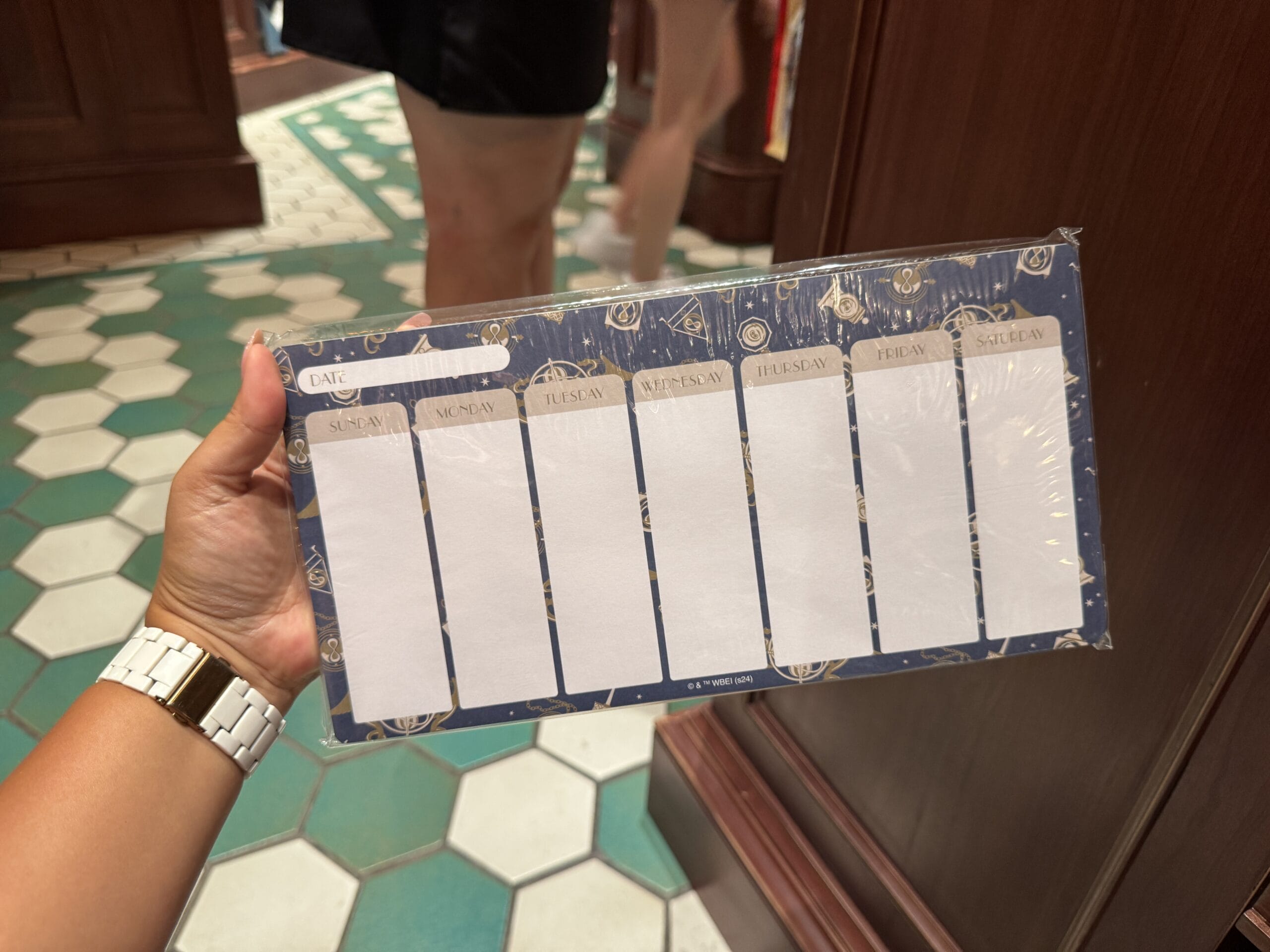 A hand holds a Wizarding World of Harry Potter merchandise planner pad with daily columns, against a tiled floor and wood wall.
