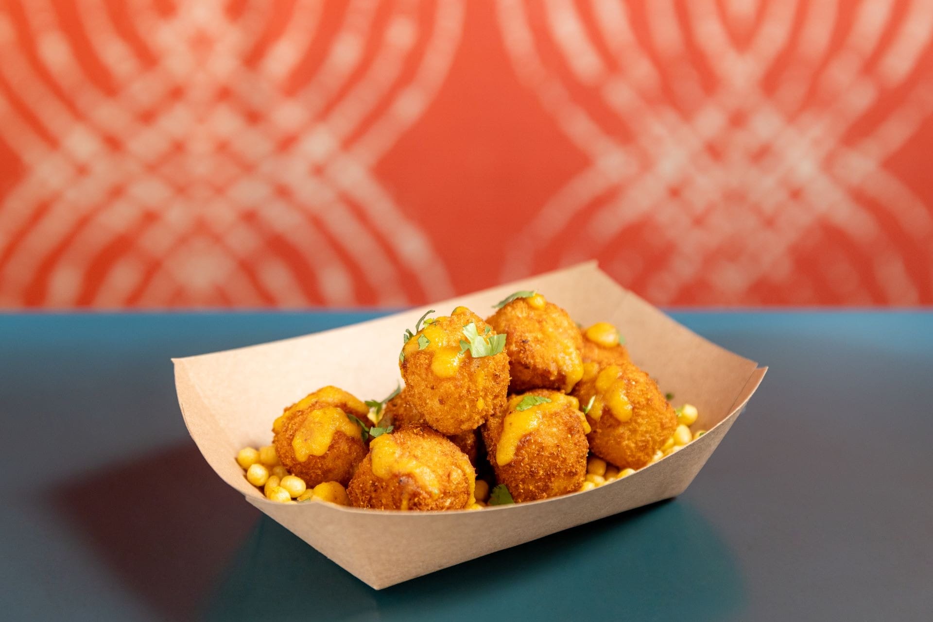 Six fried cheese balls topped with herbs in a paper tray, served over crunchy corn kernels on a blue table.