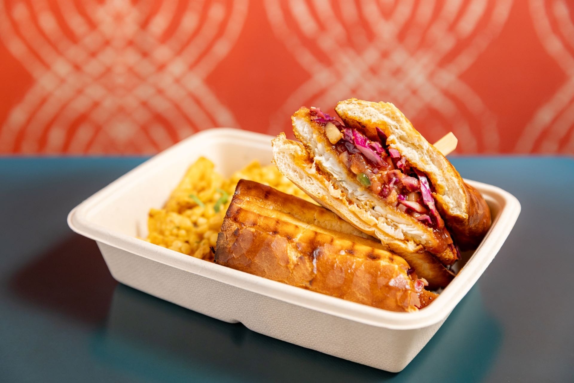 A takeout box with a sandwich filled with veggies and a side of macaroni and cheese on a table.