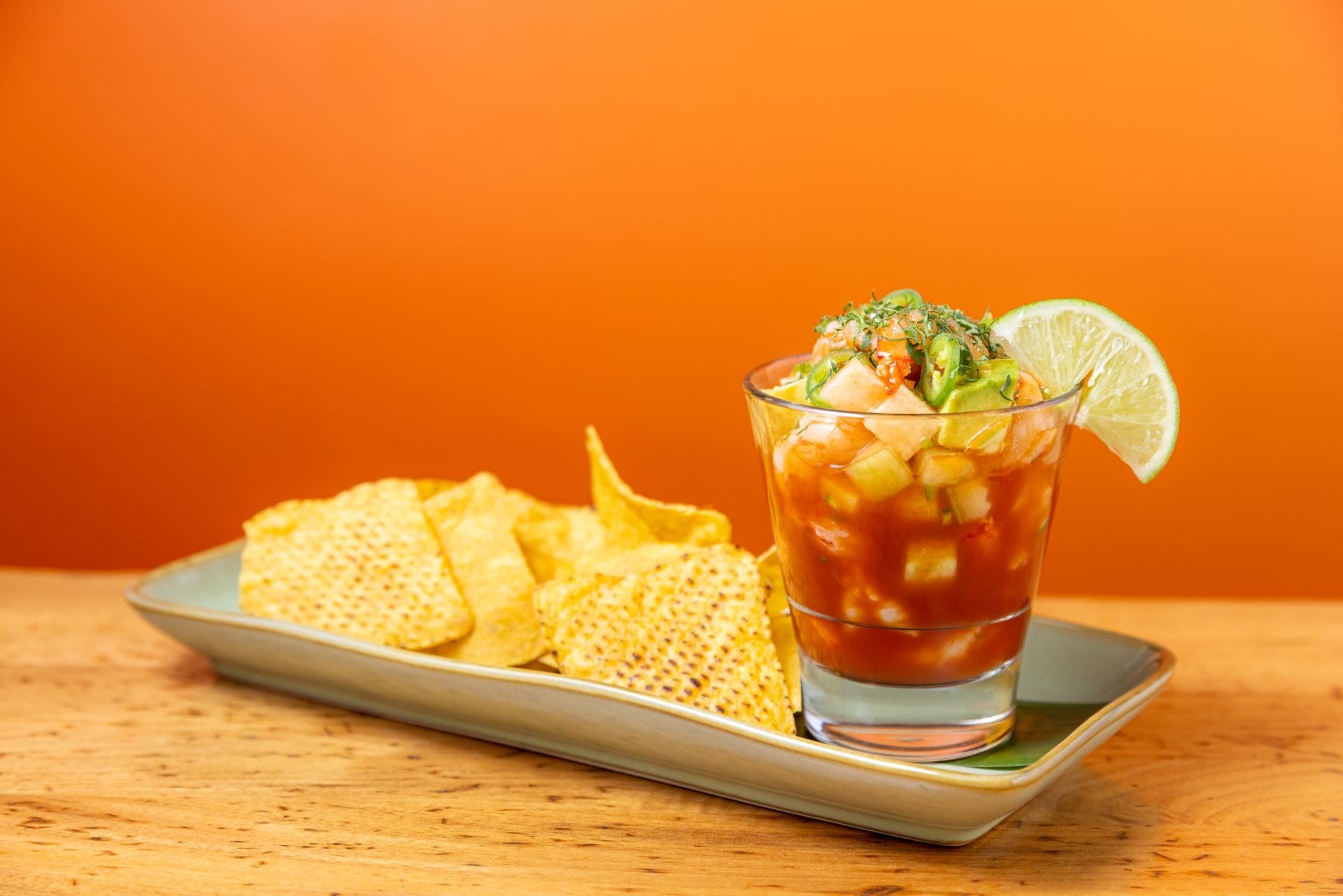 A glass of shrimp cocktail with lime, served with tortilla chips on a rectangular plate against an orange background.