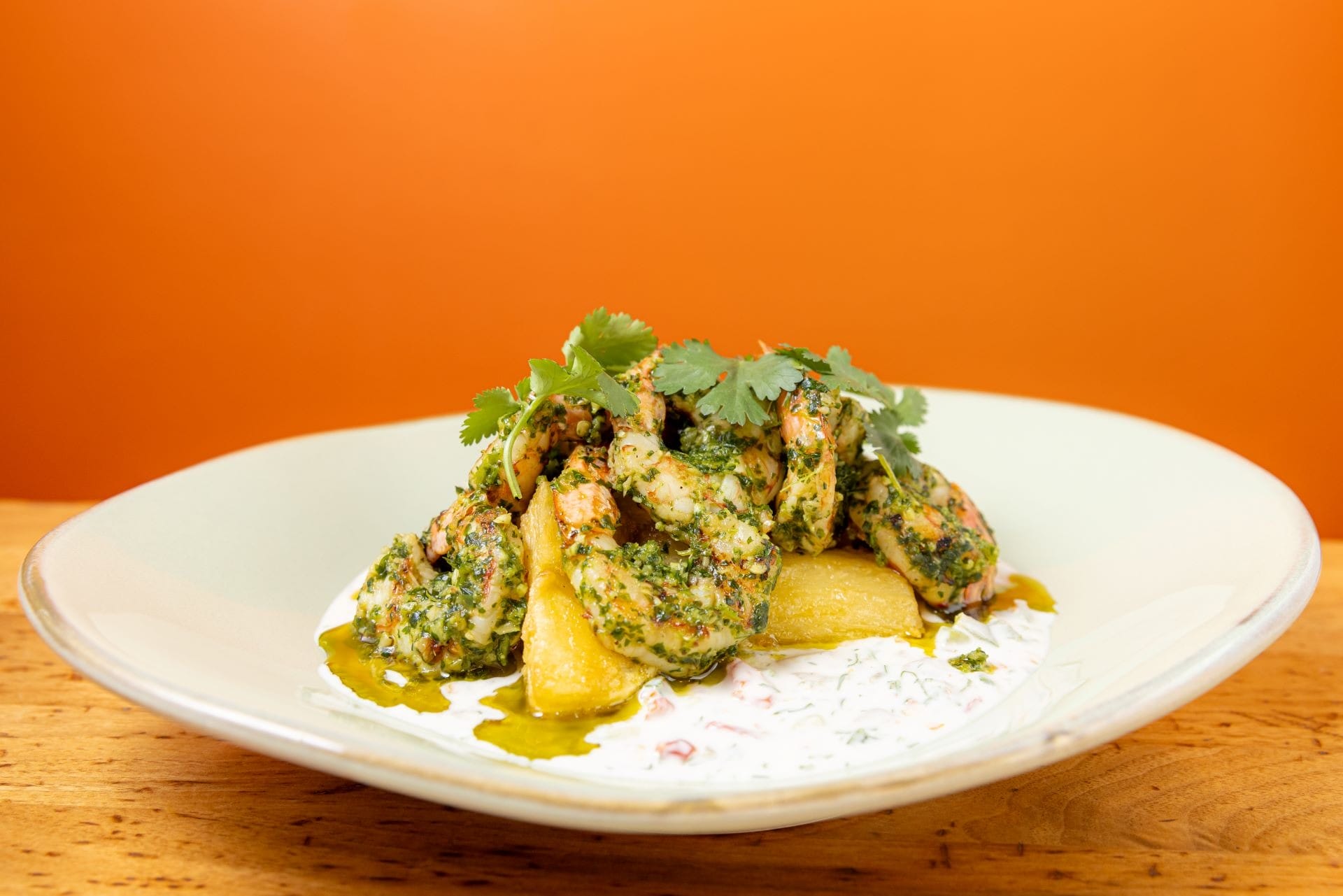 Herb-marinated shrimp and potato wedges on a white plate with yogurt sauce, garnished with cilantro, against an orange background.