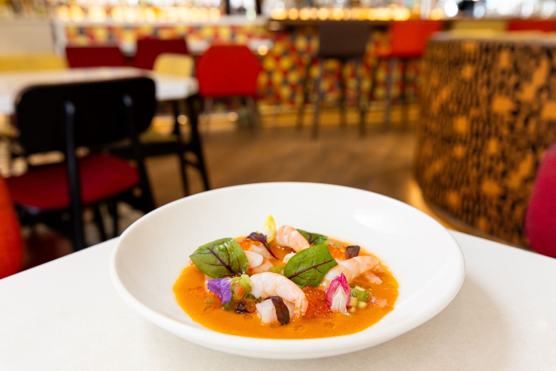 A white bowl of shrimp ceviche with colorful vegetables and sauce on a restaurant table, blurred chairs in background.