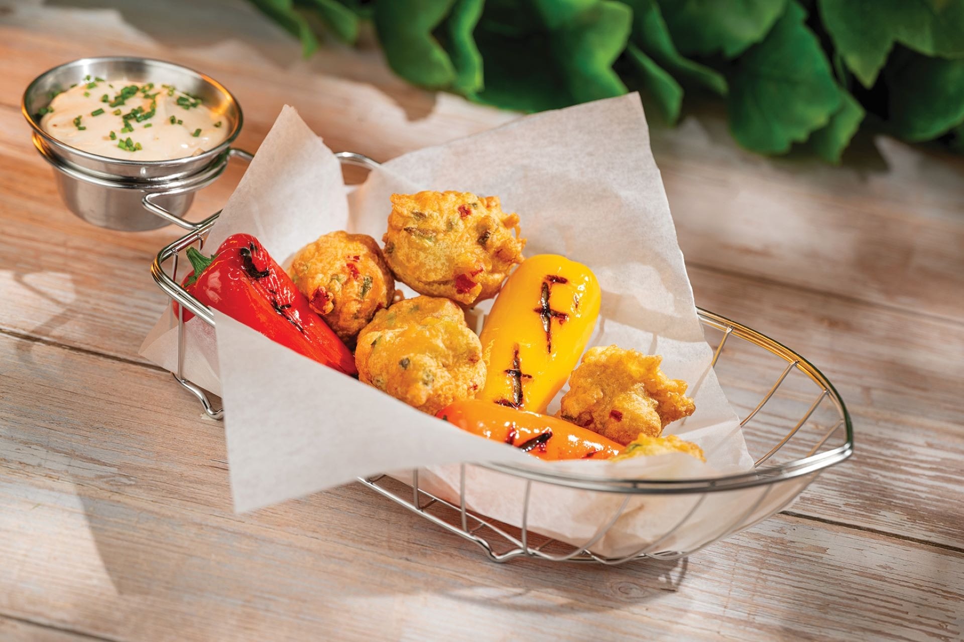 A metal basket holds fried peppers and hush puppies on parchment, with a side of dipping sauce topped with chives.