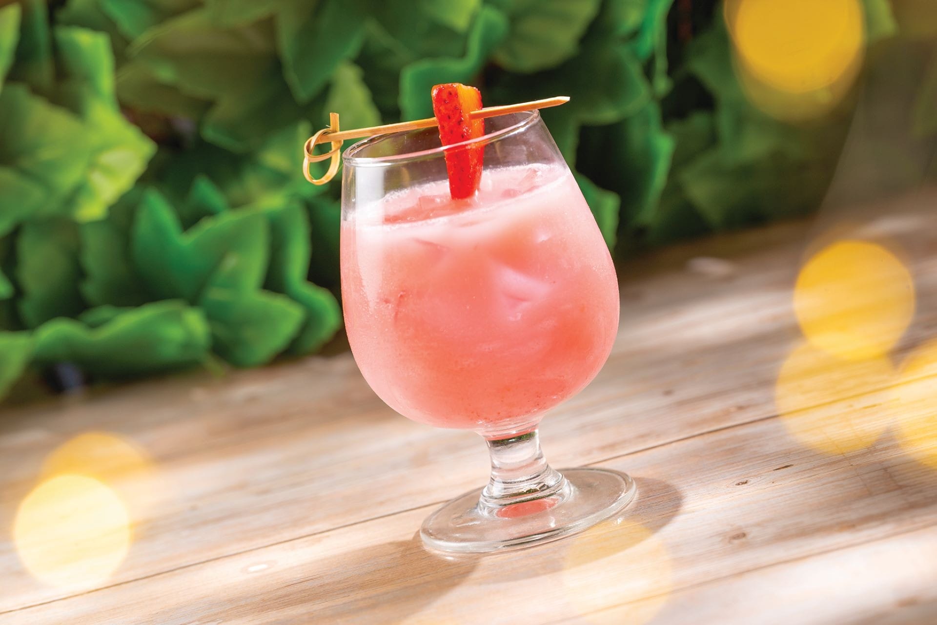 A pink iced drink in a goblet glass, garnished with a strawberry, on a wooden table with green leaves in the background.