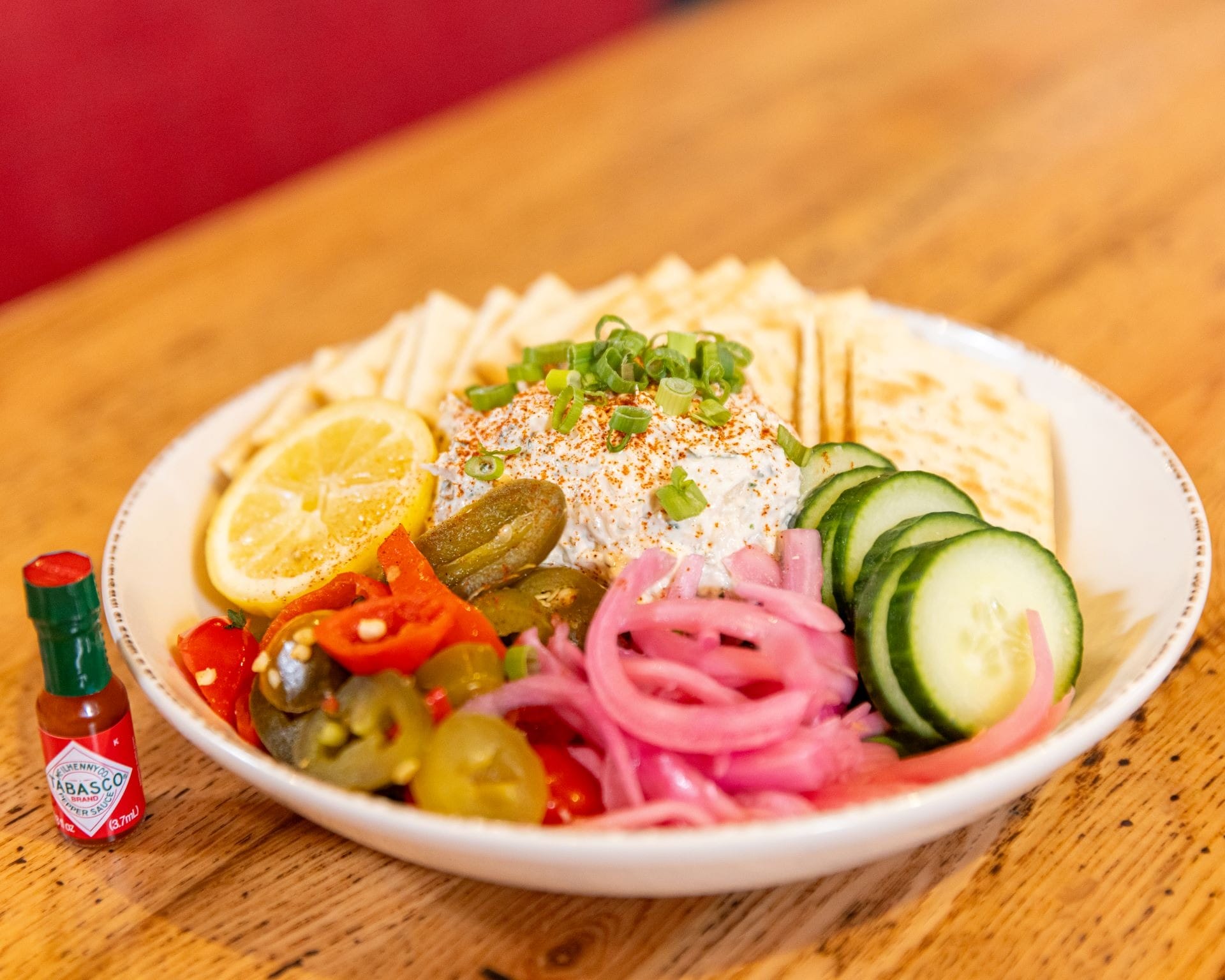 A plate with crackers, sliced cucumbers, pickled onions, peppers, a lemon slice, and a creamy dip, with hot sauce bottle.