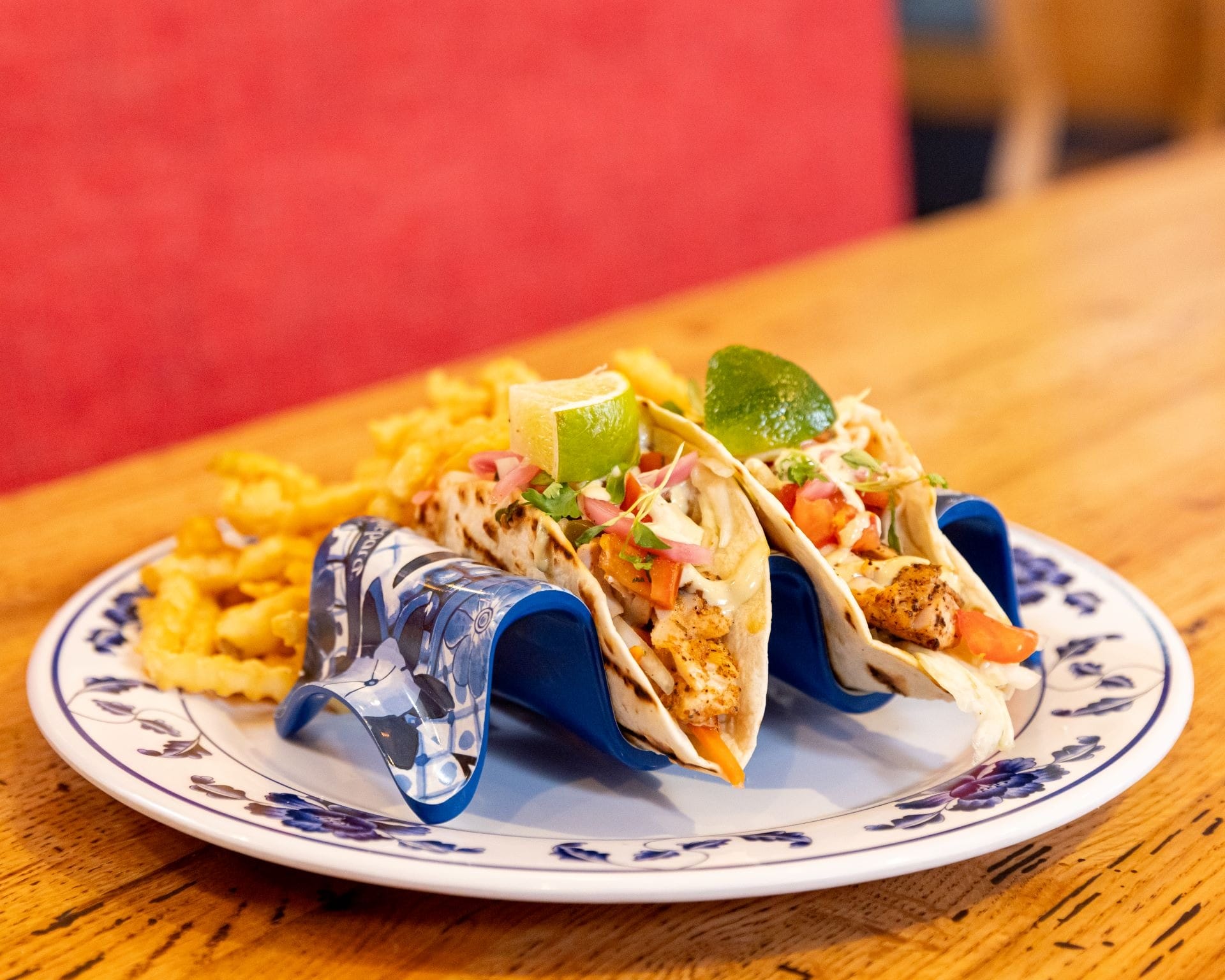 Two grilled chicken tacos with toppings and a lime wedge, served with crinkle-cut fries on a decorative plate.