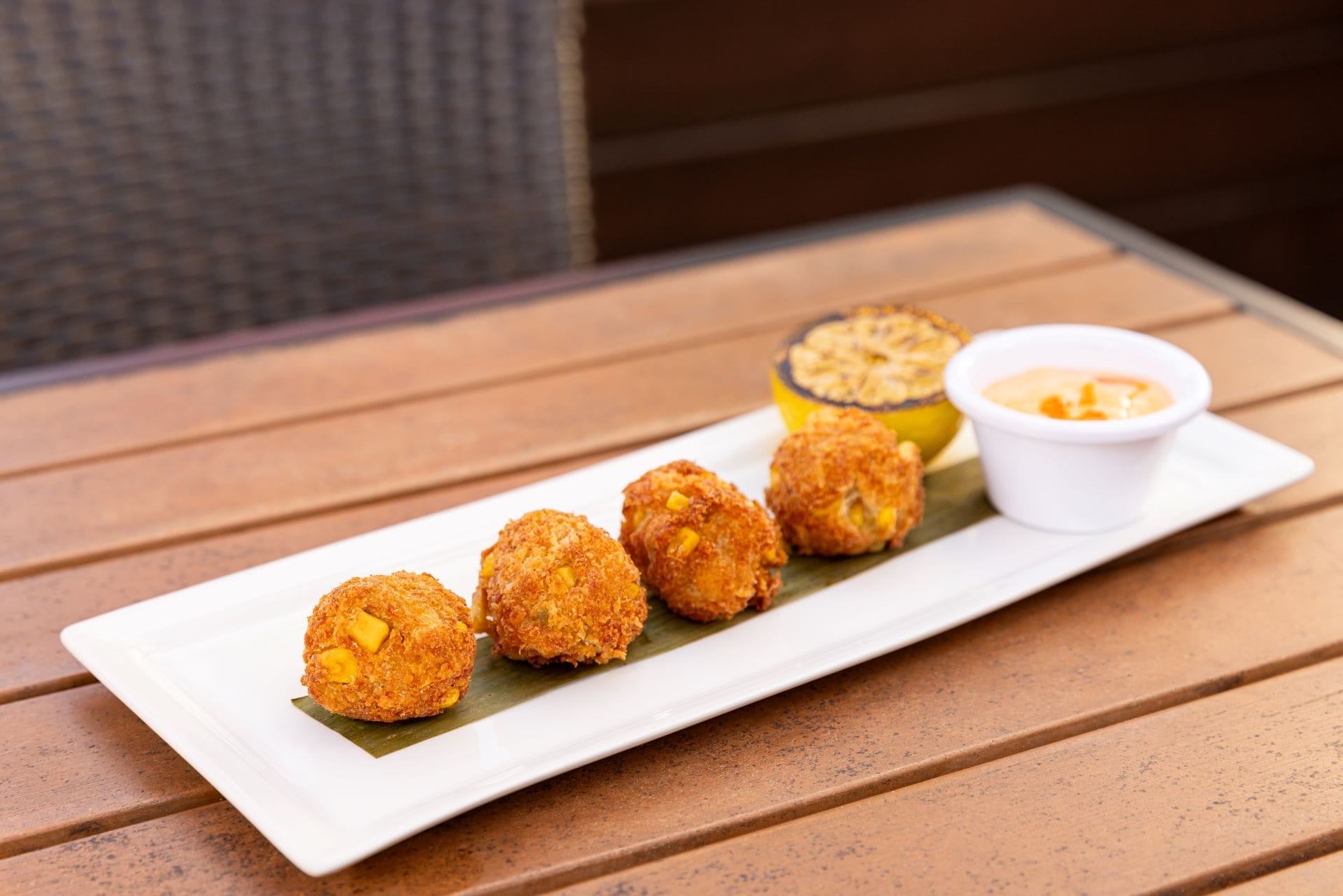Four breaded fried balls served on a rectangular plate with dipping sauce and a grilled lemon on the side.