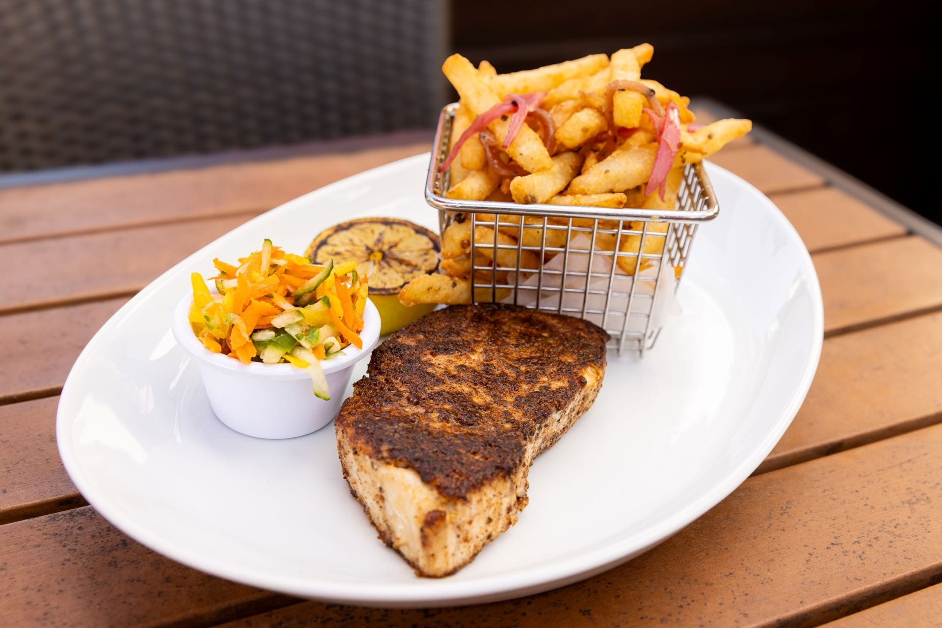 Blackened fish fillet with grilled lemon, fries in a basket, and slaw on a white plate on a wooden table.